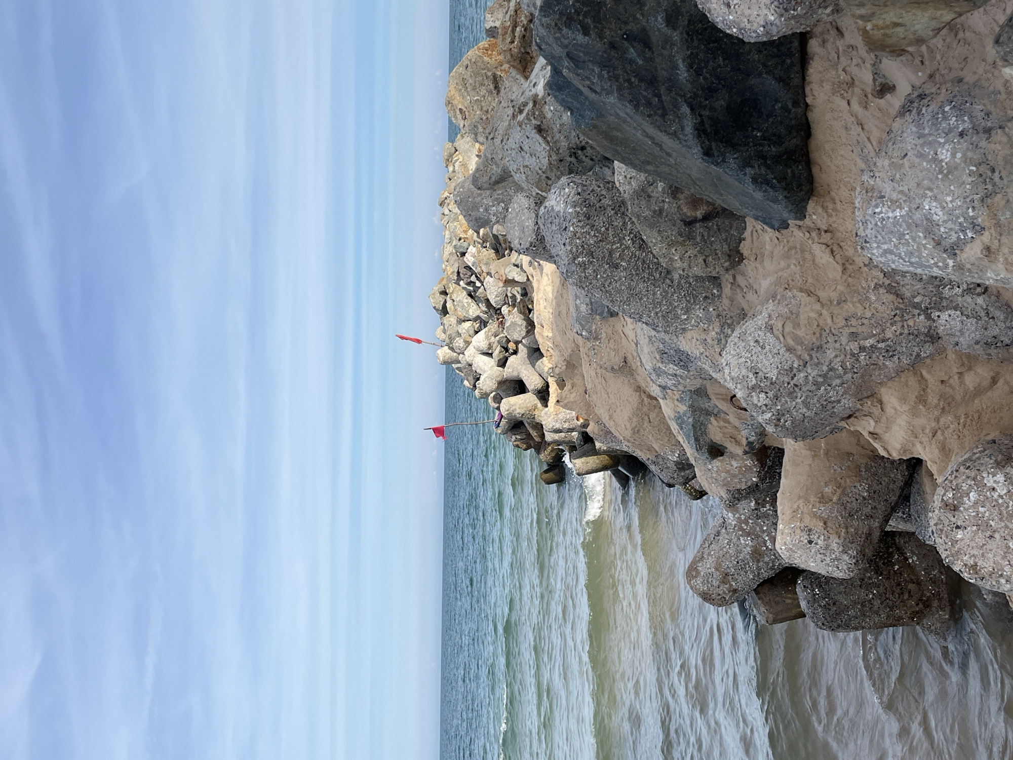 A breakwater made of large rocks and concrete tetrapods extends into the ocean under a pale blue sky with thin, wispy clouds. Two small red flags are visible on poles atop the breakwater. The ocean water is a murky green and appears choppy with small waves lapping against the rocks. The shoreline, visible in the foreground, is also composed of rocks and sand. The location is identified as Phu Thuy Ward, Vietnam. There are no people visible in the image.