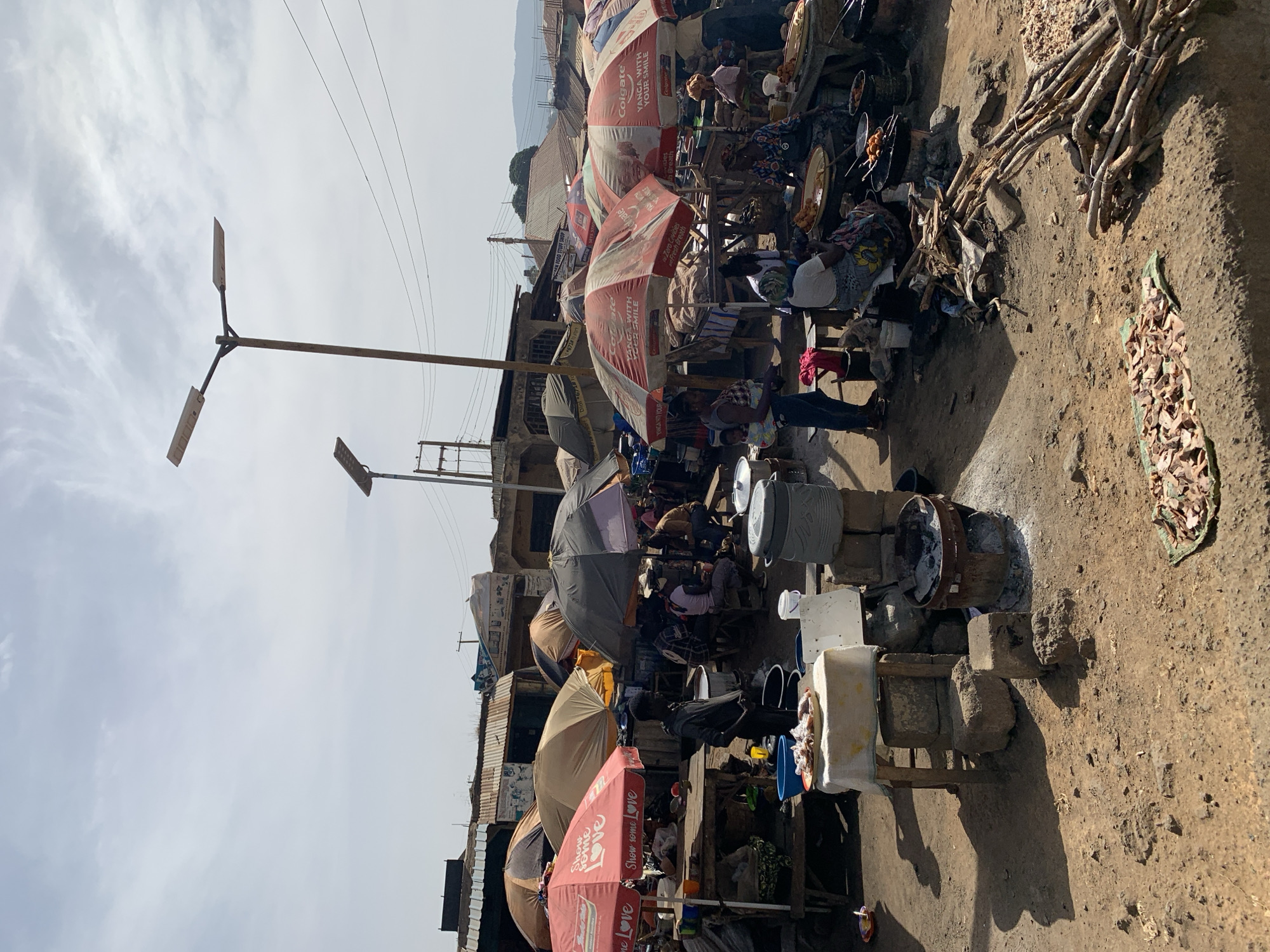 The image depicts a bustling marketplace in Rafin Par, Nigeria. The scene is filled with vendors and customers, identifiable by the array of umbrellas, stalls, and cooking equipment. Food is actively being prepared over open fires supported by stones. Cooking utensils and food are visible, including fried foods and prepared ingredients laid out on mats. Electrical poles and wiring are present, highlighting the mix of modern infrastructure amidst the traditional market setting. The ground appears uneven and dusty. Umbrellas with Colgate and other brands are visible.