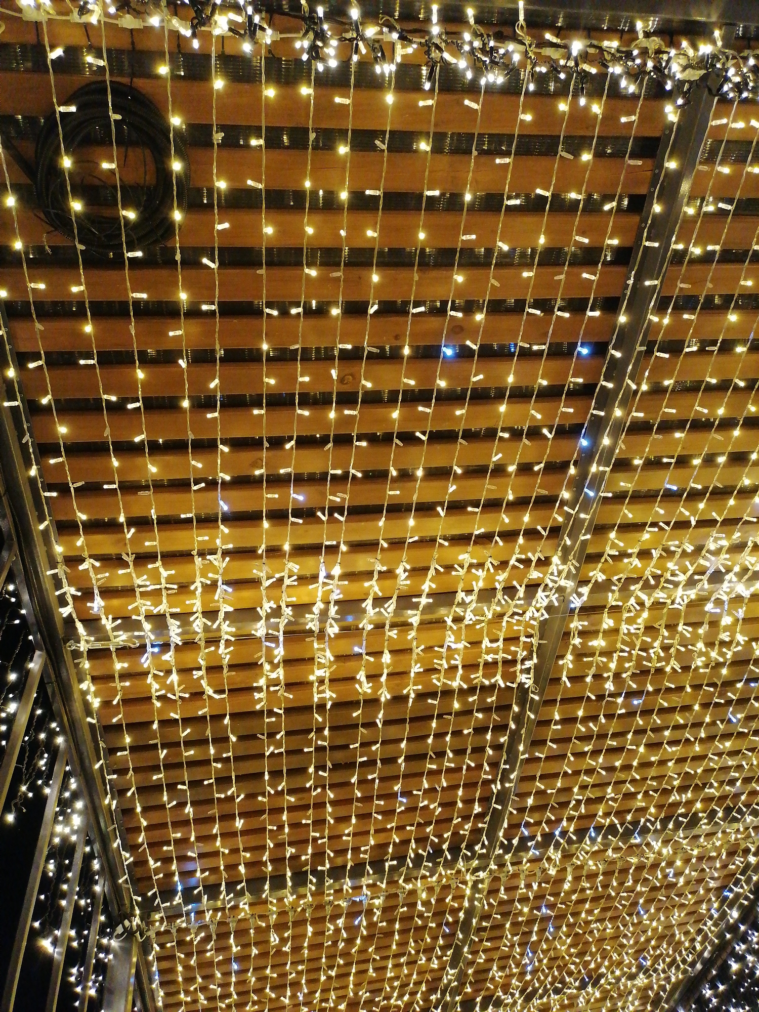 The image shows a low-angle view of a ceiling illuminated with strings of small, warm-toned lights. The ceiling consists of wooden slats, creating a grid-like pattern interspersed with the hanging lights. A coiled black cable is visible in the upper left corner. The strings of lights extend from the top to the bottom of the frame, creating a dense, glittering effect. The structure appears to be part of an outdoor space, as suggested by the partial view of additional lights and structure on the left side of the image. The location is Unknown, Unknown.