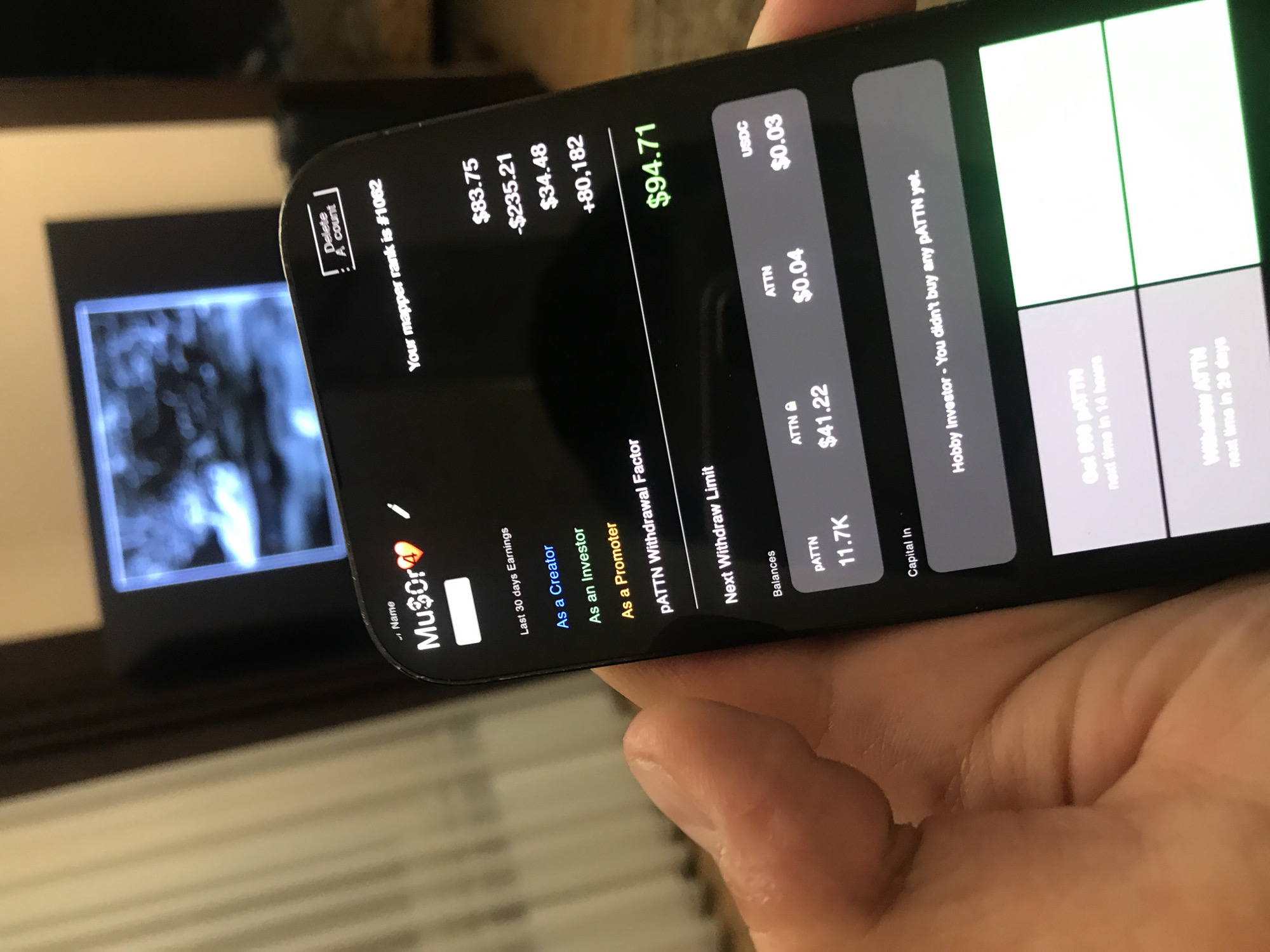 The image shows a person holding a smartphone displaying the "Musor" account screen, which is a cryptocurrency-related application. The phone screen shows a series of financial metrics and options related to the application. The account has various statistics like "Last 30 days Earnings" which includes: As a Creator ($83.75), As an Investor (-$235.21), and As a Promoter ($34.48). Other stats displayed include: PATTN Withdrawal Factor (+80,182), Next Withdraw Limit ($94.71). There are displayed balances of: PATTN (11.7K), ATTN ($41.22), ATTN ($0.04), and USDC ($0.03). The screen indicates that the user is a "Hobby Investor" who hasn't bought any pATTN yet. The user's mapper rank is #1062. A TV is visible in the background showing what appears to be a nature scene. There are no indications of Yerevan, Armenia.