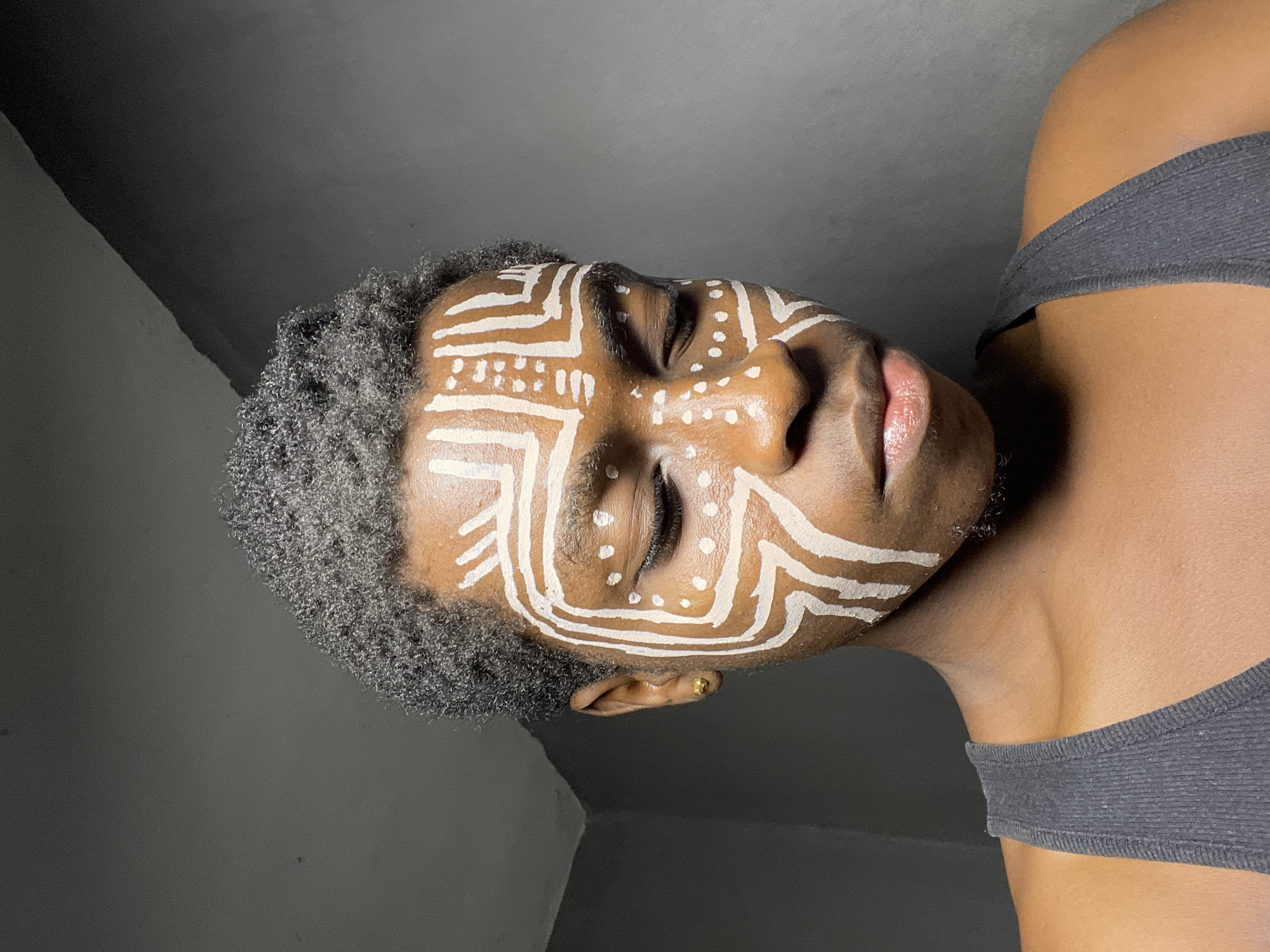 A medium shot depicts a person with dark skin and short, curly, dark hair, wearing a dark gray tank top and displaying white face paint. The design includes geometric shapes, lines, and dots. The person has their eyes closed and is angled downward. A gold earring is visible on their left ear. The background consists of a gray wall. There is no information available to determine if the image was taken in Nkpor, Nigeria.