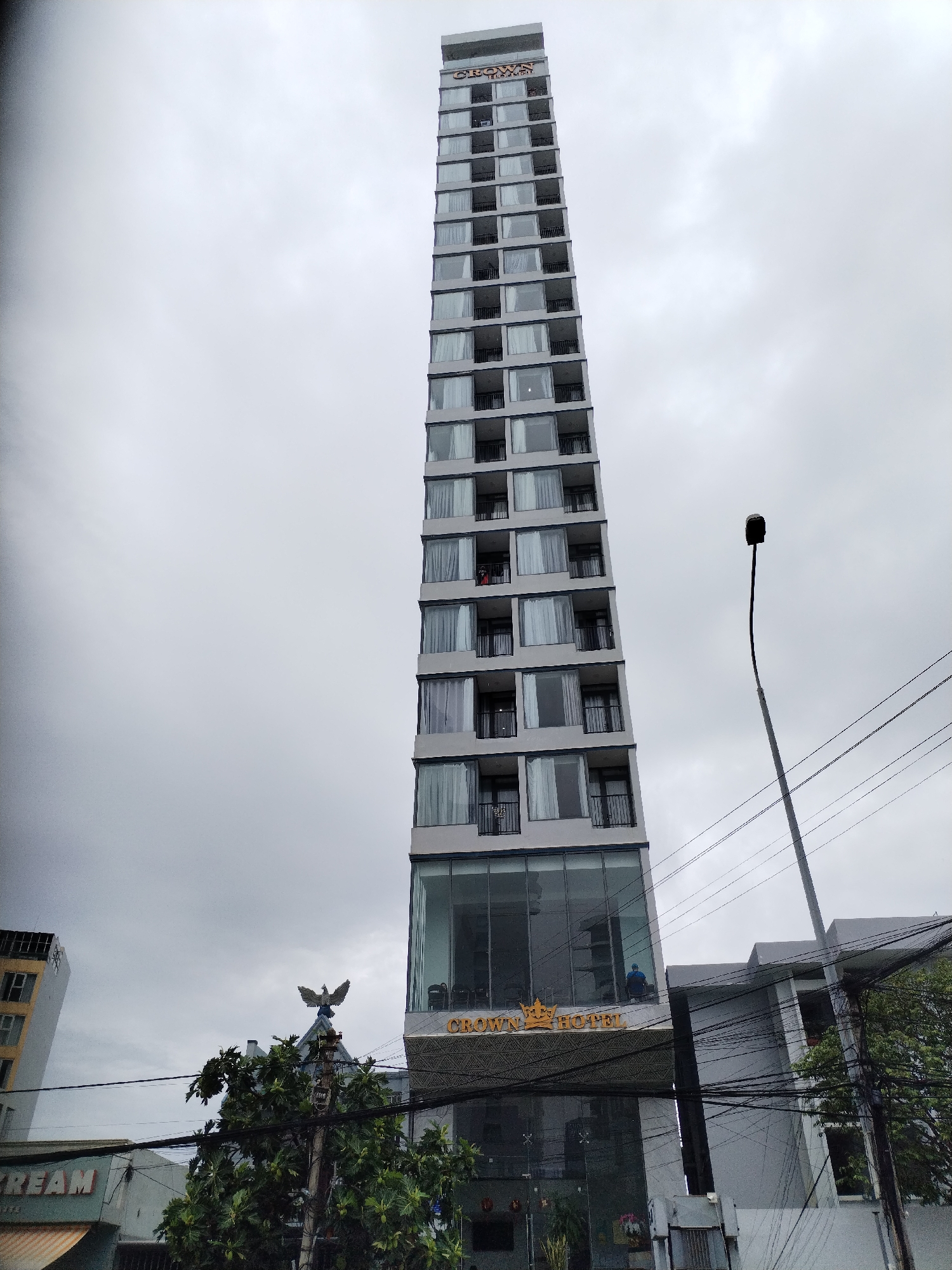 A tall, narrow, modern building dominates the center of the frame, stretching upwards under an overcast sky. This multi-story structure features numerous windows and small balconies on each floor, with a light grey facade and dark window frames and railings.
Near the top of the building, partial golden text spelling "CROWN" is visible. At the base, a large glass-fronted lobby area displays more golden lettering that reads "CROWN HOTEL." A person in a blue shirt can be faintly seen inside the glass lobby. On an upper floor, a person wearing red is visible on one of the balconies.
To the right of the main building, a tall, slender street lamp with a dark, curved head extends into the frame. A dense tangle of black overhead power lines crisscrosses the midground, running between structures and over a leafy green tree situated in the lower left. On the roof of a smaller building to the left, a golden statue of an eagle-like bird with outstretched wings is prominently displayed. Another building in the lower-left foreground has partial white text, "REAM," visible on its facade.
The scene depicts an urban environment during what appears to be an overcast day, indicated by the uniformly gray sky. The dense infrastructure, including the power lines and varied architecture, is consistent with an urban setting in Nha Trang Ward, Vietnam.