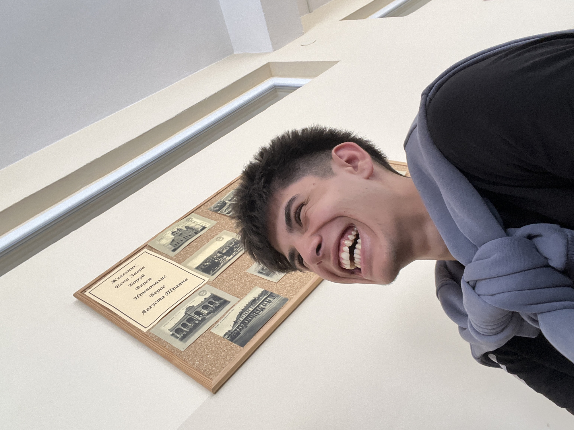 A low-angle, medium shot captures a young man with dark, spiky hair laughing. He is wearing a black shirt and a light blue sweater tied around his neck and shoulders. Behind him, a corkboard displays several vintage black and white photographs of buildings. A sheet of paper with Cyrillic text is pinned to the board, and the text on the paper includes "Железник," "Ески Заара," and "Стара Загора," indicating the location is in or related to Stara Zagora, Bulgaria. The background consists of a white wall and a section of a ceiling with a light fixture, suggesting an indoor setting, likely a school or public building.