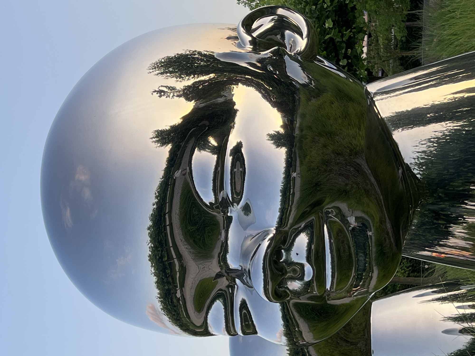 The image is a close-up shot of a large, reflective sculpture of a baby's head. The sculpture is made of polished metal, mirroring the surrounding environment. The reflection shows a park-like setting with green grass, trees, and a clear blue sky with some clouds. The setting sun casts a warm glow, visible in the reflections of the sky and trees. In the distance, there are hints of people walking and a path. The sculpture is located in the City of Krasnodar, Russia, specifically in the Public Garden of Krasnodar.