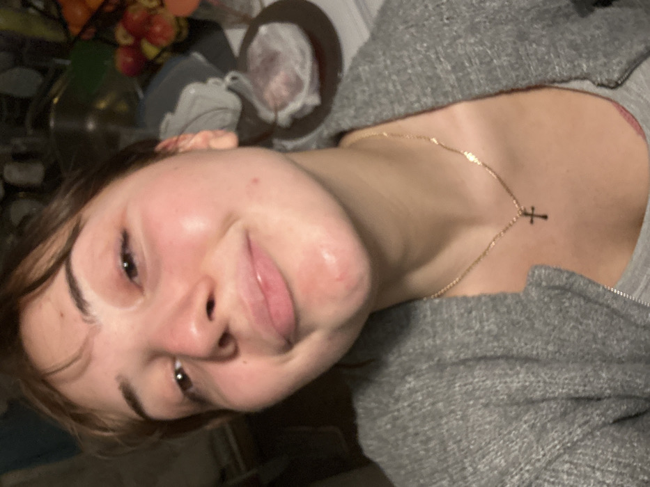 A young woman with fair skin, dark eyes, and rosy cheeks smiles slightly at the camera. She is wearing a gray hooded jacket and a gold necklace with a small cross pendant. Her hair is pulled back. Behind her, a kitchen countertop is visible with fruit in a bowl and other miscellaneous items. The image has a slightly rotated perspective.