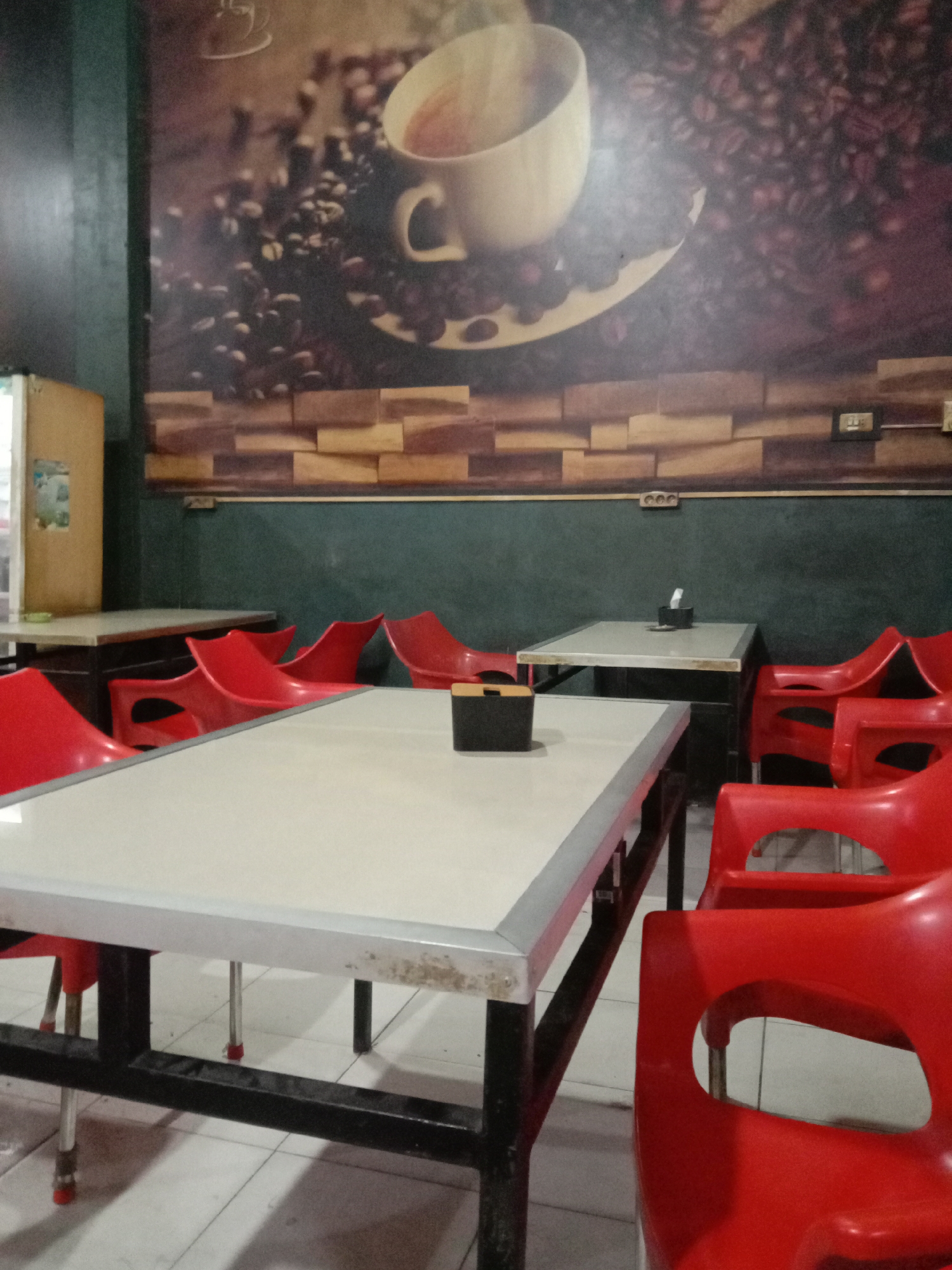 The image displays the interior of an empty coffee shop or cafe, likely located in Lhokseumawe, Indonesia. The setting appears casual and functional, awaiting customers.
In the foreground and central midground, a rectangular table with a light-colored, possibly white or off-white, top and a sturdy dark metal frame dominates the view. A dark, possibly black, tissue holder is placed on this table. Surrounding it and extending into the background are multiple identical red plastic chairs, some arranged neatly at tables, others pushed in. Another similar table is visible further back on the right side of the room.
The back wall features a large mural depicting a white coffee cup brimming with a hot beverage, surrounded by numerous dark coffee beans. Below this coffee-themed mural, the wall is painted a dark greenish-grey, punctuated by a horizontal band of brown, wood-effect paneling or tiling. Several electrical outlets are visible on this lower wall section. To the far left, a light-colored cabinet or service counter is partially visible. The floor is covered with light-colored tiles.
The absence of people suggests the image was taken during off-peak hours or when the establishment was closed. No discernible text is visible within the frame. The decor, particularly the mural, clearly communicates the venue's purpose as a place for coffee or refreshments.