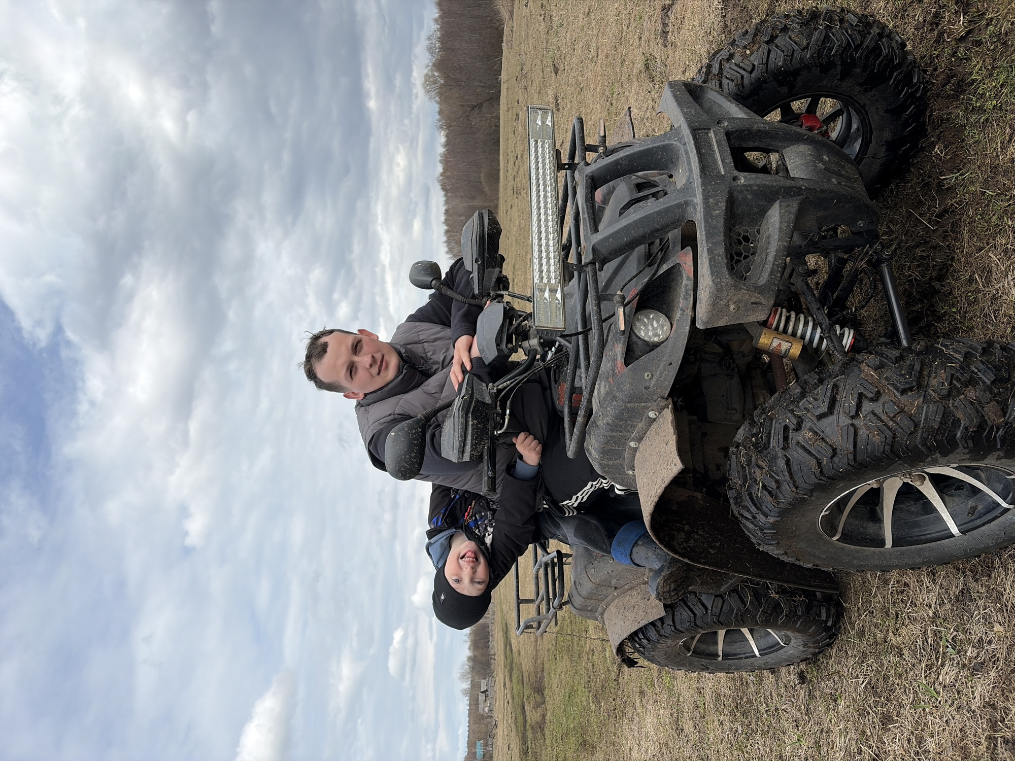 Two individuals, likely a father and son, are positioned on a black ATV in a field near Кемуль, Russia. The ATV's tires appear muddy. The sky is overcast. The father sits on the ATV with the child in front of him. A light bar is visible on the ATV. Trees are visible in the background.