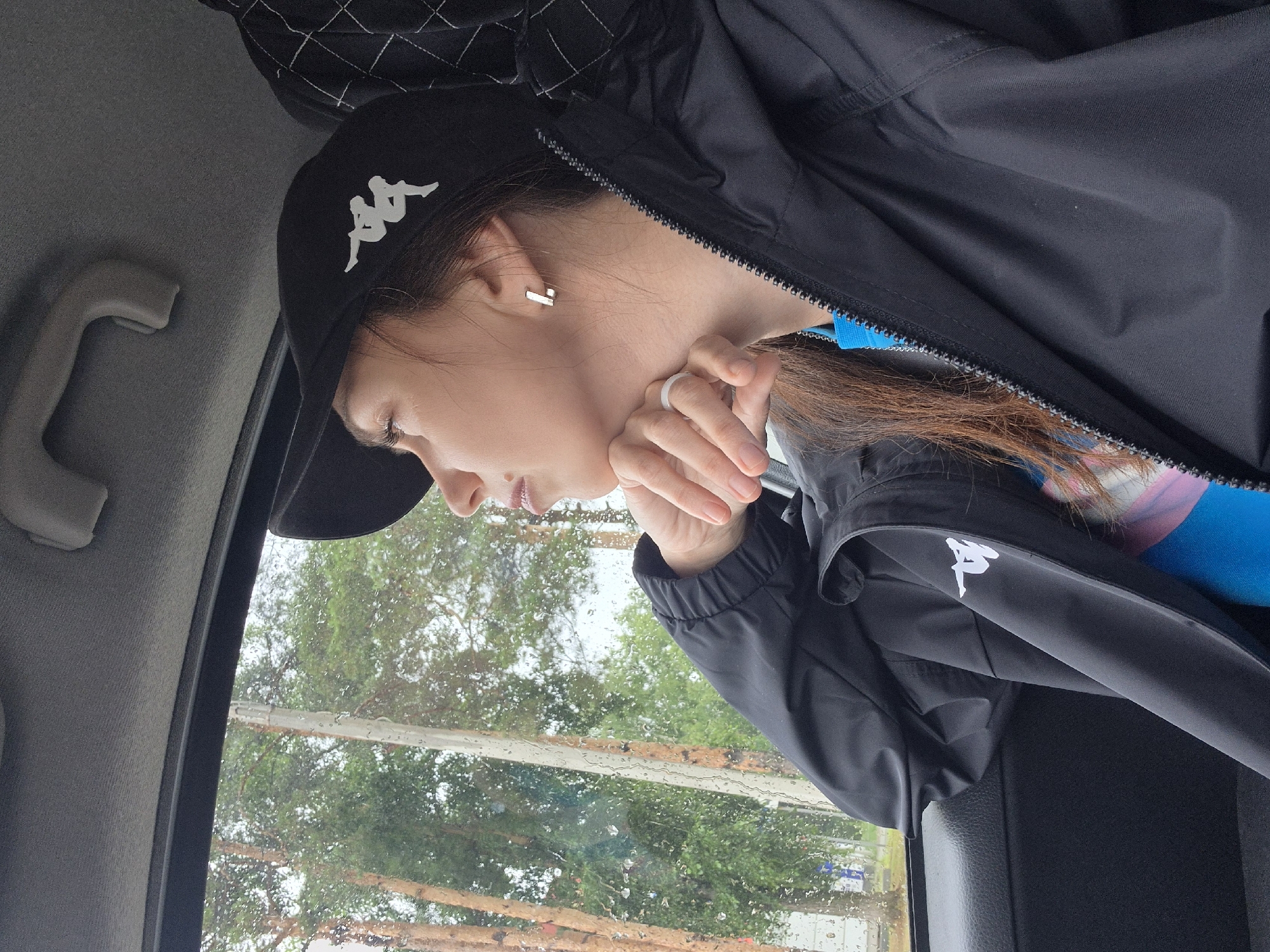 A medium shot shows a young woman wearing a black baseball cap with a white logo, a black jacket with a white logo, and a blue shirt. She is in the passenger seat of a car, looking out the window at a wooded area with trees. The window has raindrops on it. Her left hand rests on her chin. She is wearing a silver ring on her left ring finger and a silver rectangular earring in her left ear. The car interior is dark gray.