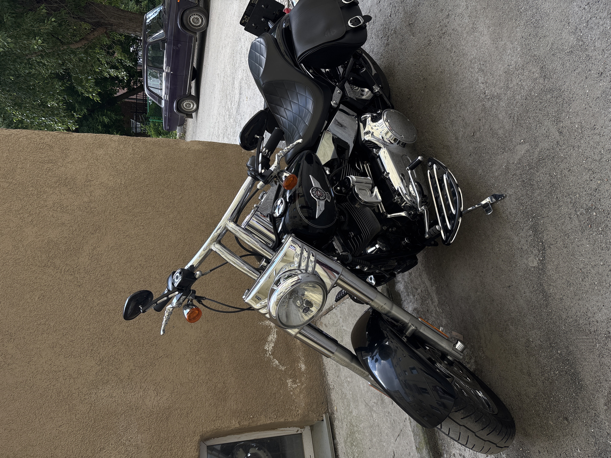A black Harley-Davidson motorcycle is parked on a paved surface next to a textured, light brown wall. The motorcycle has chrome accents, including the handlebars, front forks, and engine components. It features a diamond-stitched black leather seat and black leather saddlebags on either side. In the background, a dark purple sedan is parked, and a large tree with green leaves is visible. The scene appears to be an outdoor urban setting, possibly in a courtyard or alley. The image is taken from a slightly low angle, focusing on the motorcycle. The location is identified as Novosibirsk, Russia.
