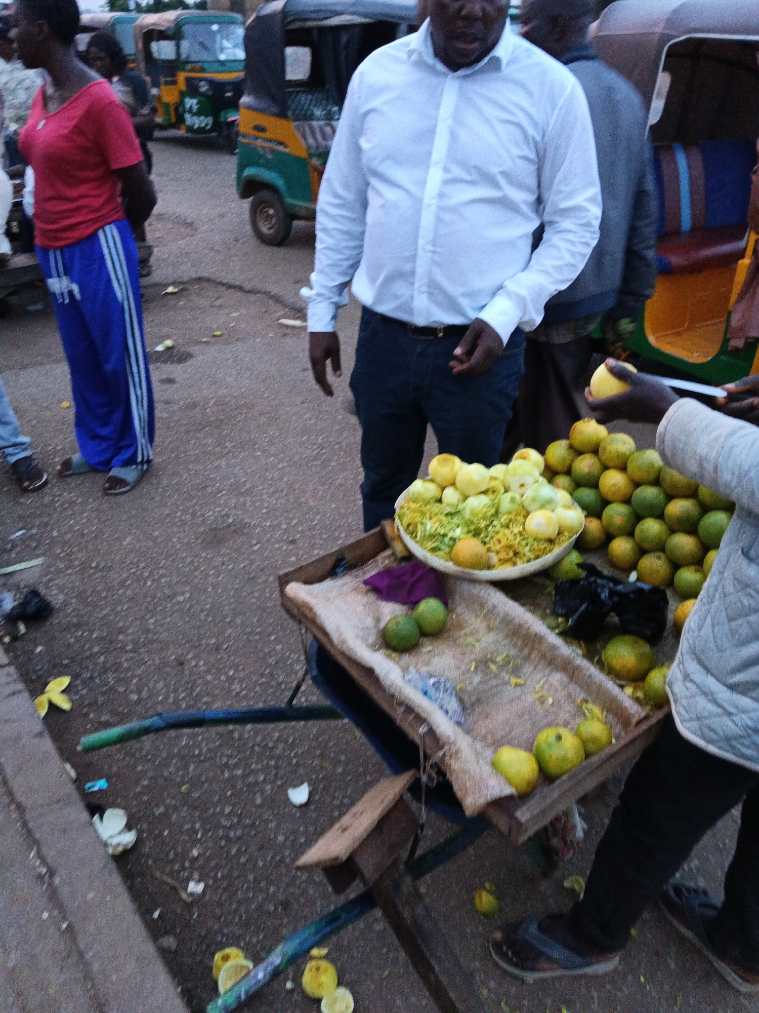 In a street scene in Bukuru, Nigeria, an outdoor market stall displays a large bowl of sliced citrus fruits, possibly oranges, alongside piles of whole fruits. The vendor, dressed in a light-colored jacket and dark pants, is in the process of holding up a single piece of fruit. The stall itself is a wooden cart with a fabric covering. In the background, several auto-rickshaws are visible, suggesting a busy transportation hub. A woman in a red shirt and blue pants stands to the left, facing away from the camera. The ground is paved with asphalt and scattered with debris, including fruit peels and wrappers. The overall impression is of a vibrant, everyday market environment.