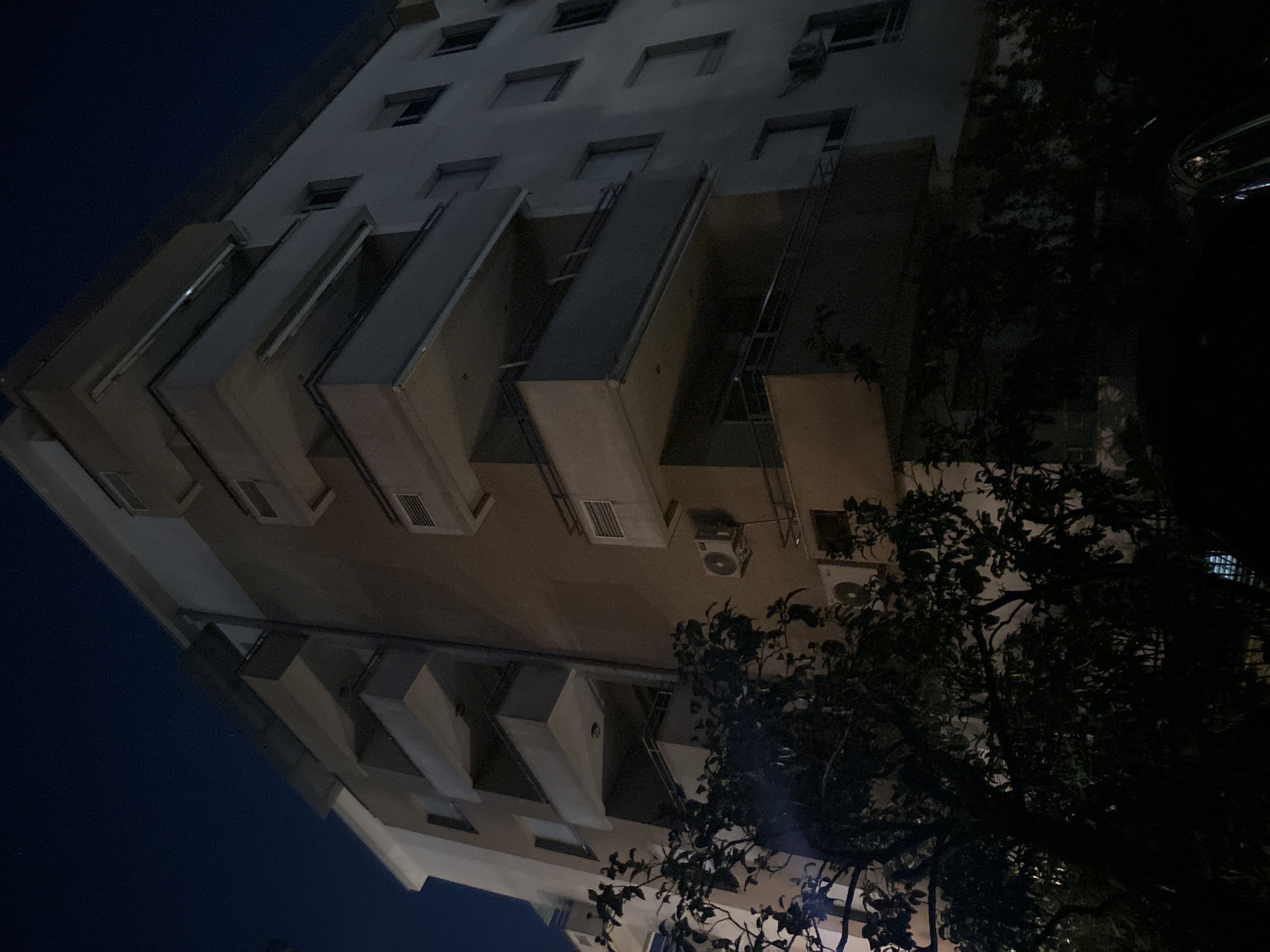 The image shows a low-angle shot of an apartment building at night, possibly in Budva, Montenegro, given the context of the prompt. The building is multi-story with several balconies. The sky is dark blue and appears clear. In the foreground, dark foliage from trees partially obscures the lower portion of the building and a hint of a car roof is visible at the bottom right. The building's facade is a mix of light gray and brown sections. Several windows are visible, some with what appear to be closed shutters. Air conditioning units are mounted on some of the exterior walls. The overall scene is dark, suggesting it was taken after sunset with minimal artificial lighting contributing to the illumination of the building. There are no people or animals visible in the frame.