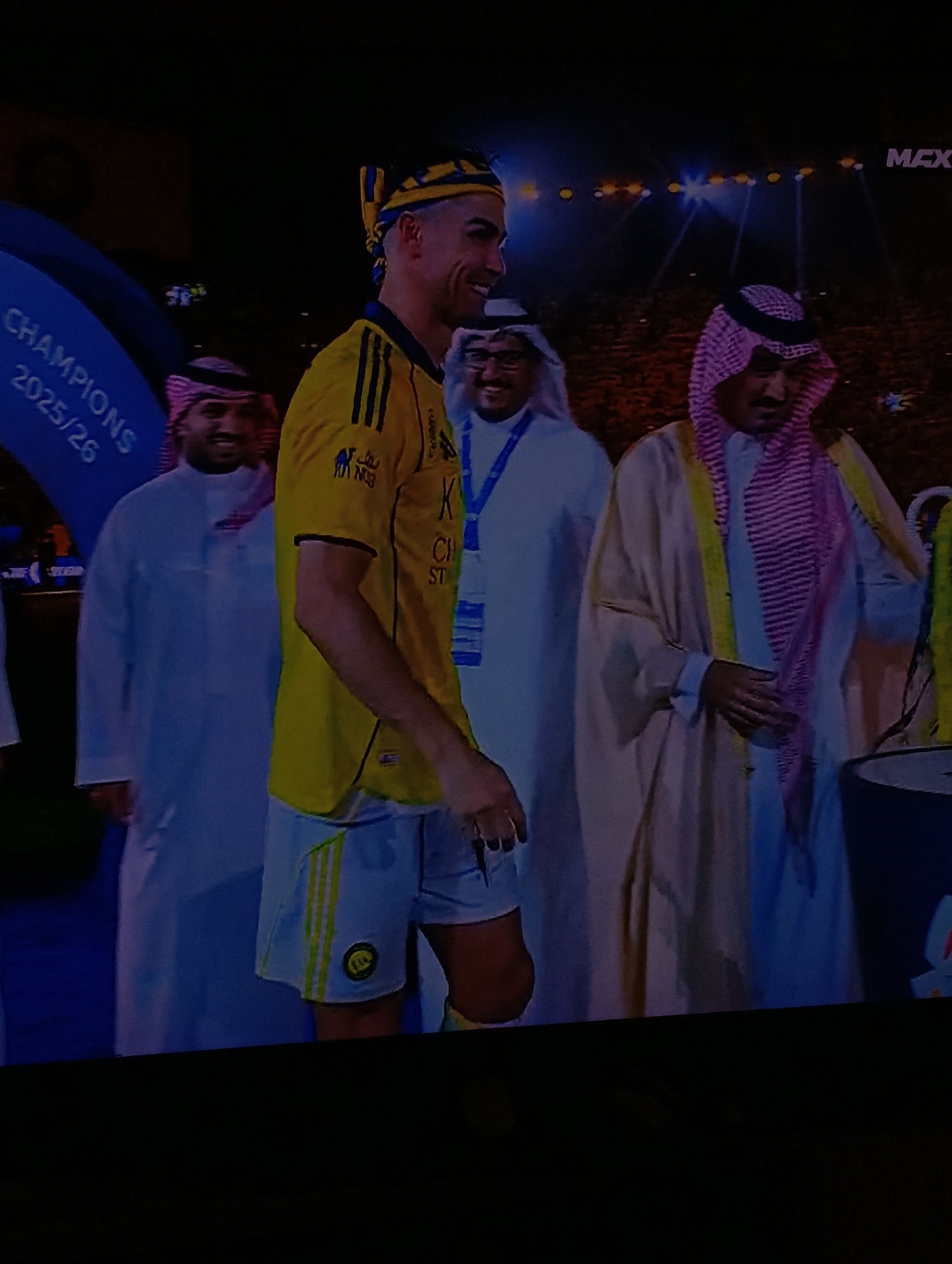 The image shows Cristiano Ronaldo, in a yellow and black Al Nassr soccer jersey and shorts, wearing a yellow and blue headband, standing with three men in traditional Saudi Arabian clothing. The setting appears to be a stage or platform, possibly after a soccer event. A banner partially visible to the left reads "CHAMPIONS 2025/26." The background suggests a crowd and bright lighting. The image was not taken in Plovdiv, Bulgaria.