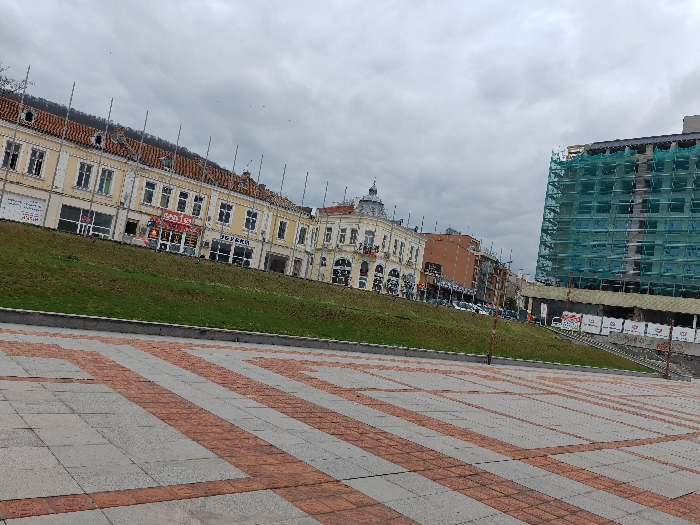 The image depicts a static urban scene in central Shumen, Bulgaria, specifically a public square area adjacent to a commercial street. The sky is uniformly overcast and light grey.
In the foreground, a large paved plaza is visible, composed of light grey square tiles interspersed with reddish-brown rectangular paving stones arranged in perpendicular stripes and a grid pattern.
Rising steeply from the edge of this plaza, separated by a dark grey curb, is a manicured grassy embankment. Several slender, dark vertical poles are spaced along this slope.
In the background, on an elevated plane behind the grass, a row of multi-story buildings with light yellow facades and red tiled roofs extends horizontally to the left. These structures feature numerous windows and ground-level commercial establishments displaying signage, including "ОБУВКИ" (shoes) and "САЯ." One building presents a more ornate, classical architectural style with a small, rounded dome. Further to the right in the background, a tall, multi-story building under construction is entirely encased in bright green scaffolding netting. Below it, a lower, flat-roofed structure with white banners is visible. A street with parked vehicles runs between the grassy embankment and these background buildings.
No individuals are visible within the frame. The overall impression is one of an active city under a subdued sky, with both established architecture and ongoing development present.