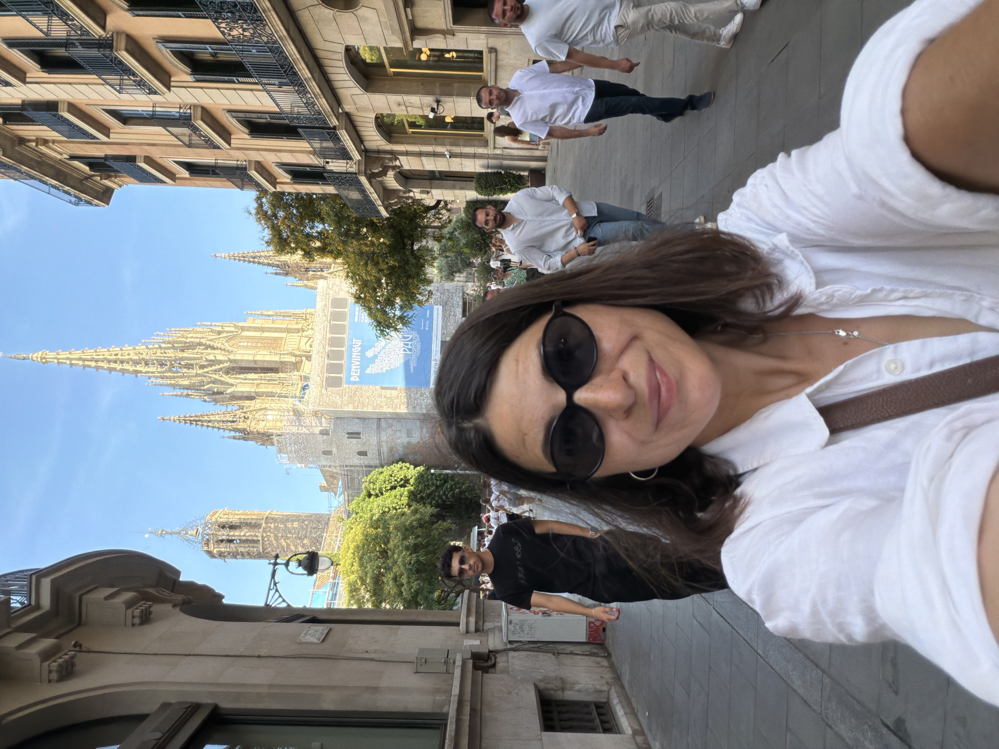 A woman wearing sunglasses and a white shirt takes a selfie in a narrow street in Barcelona, Spain. Behind her, a man in a black t-shirt walks away from the camera. In the background, people are walking on the sidewalk, and a large Gothic cathedral with spires is visible, along with a banner that reads "BENVINGUT PALI". The street is lined with buildings with balconies and windows, and trees can be seen. The sky is blue and clear, suggesting a sunny day.