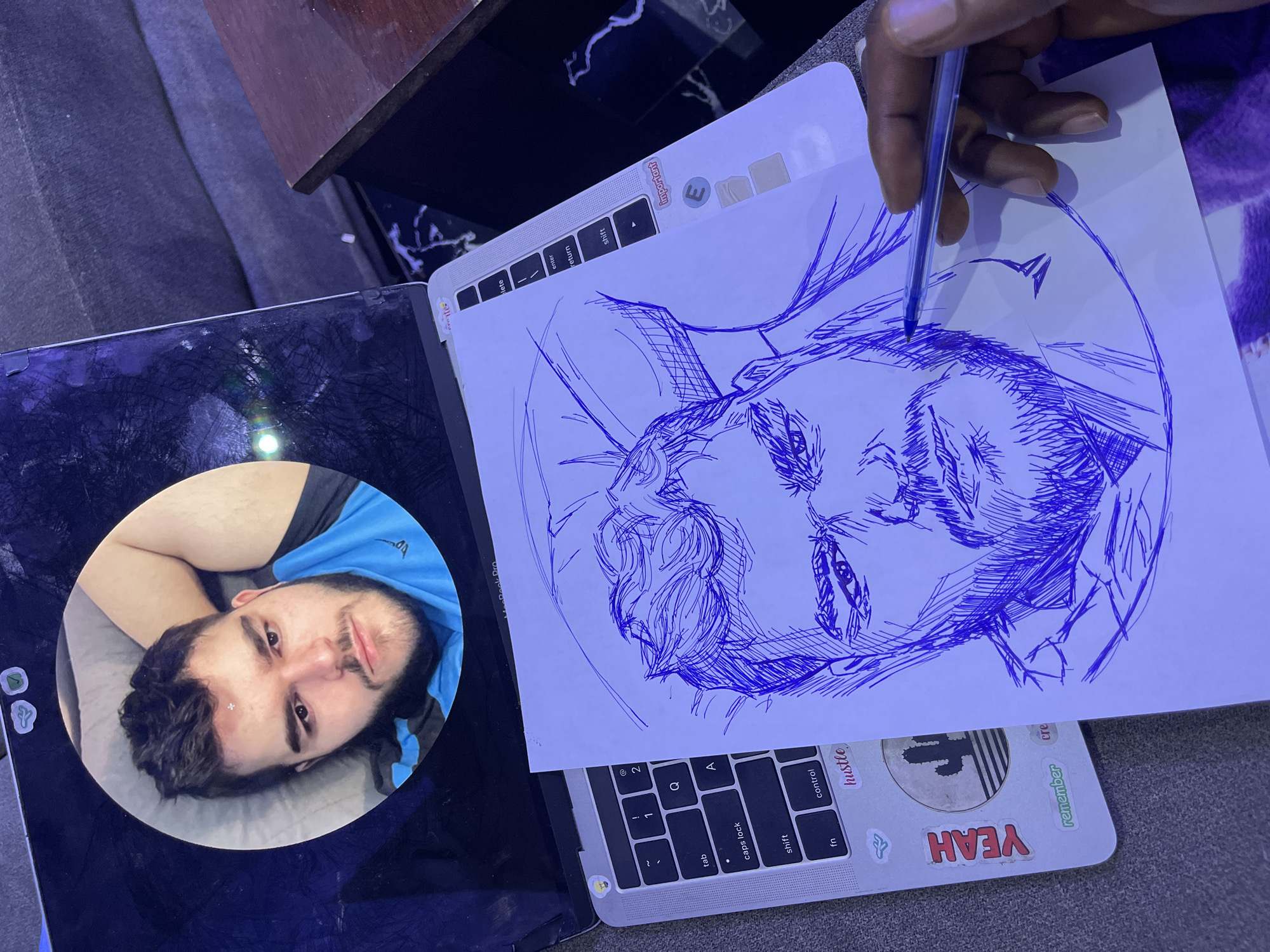 A low-angle, eye-level shot shows a person's hands drawing on a piece of paper placed on top of an open laptop. The laptop displays a circular profile picture of a man with dark hair, a beard, and a blue shirt. The drawing on the paper is a blue ink sketch of the same man. The laptop is a MacBook Pro, indicated by the text on the screen. The keyboard has various keys, including 'tab', 'caps lock', 'shift', 'fn', 'control', 'Q', and 'A'. There are stickers on the laptop, including one with the word "YEAH" and another with a cactus. The person is using a blue ballpoint pen to draw. The scene is indoors, with a dark-colored couch visible in the background and a wooden table to the right. The lighting is dim and possibly artificial. The location is not specified as being in Abuja, Nigeria.
