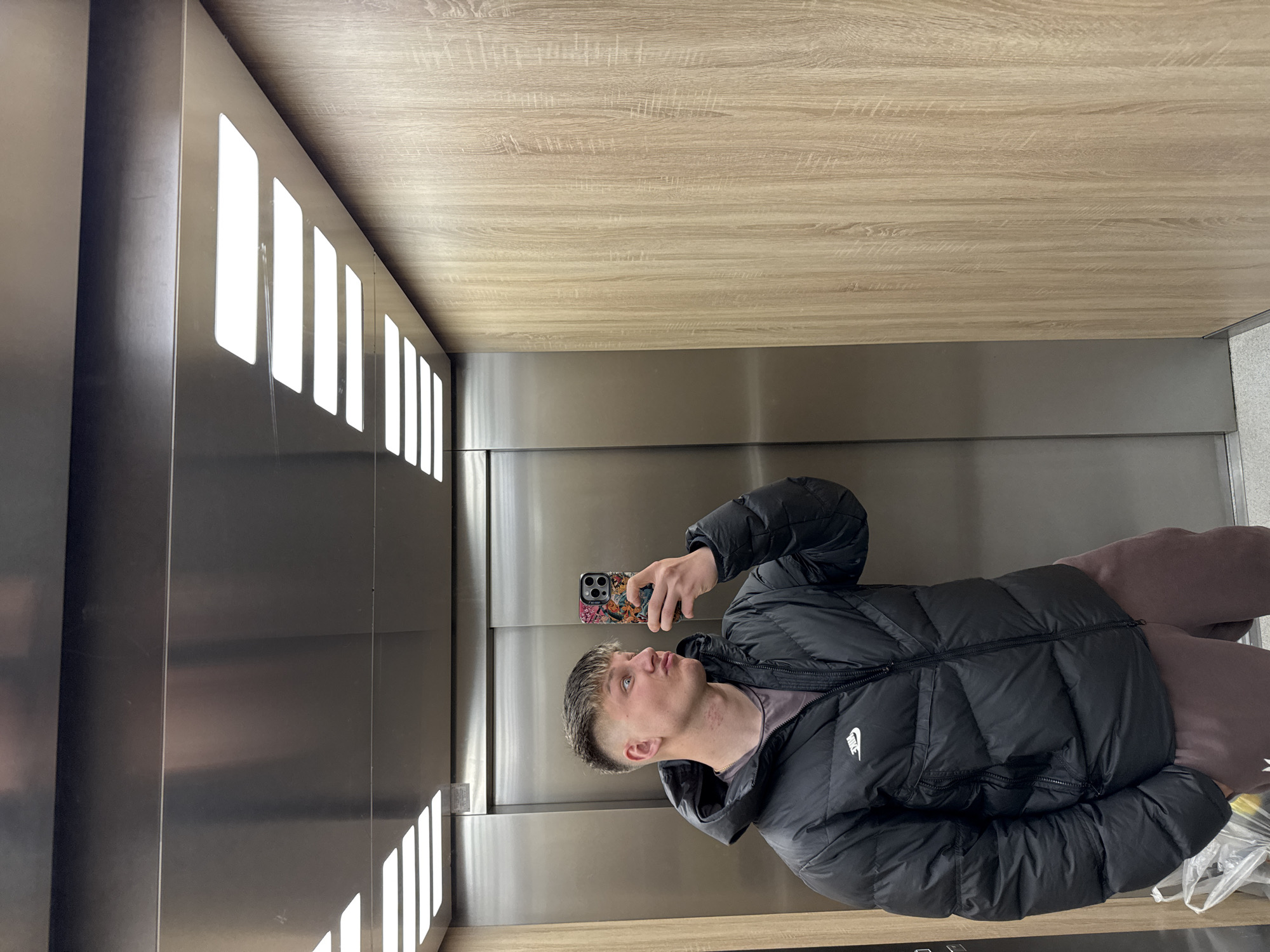 A young man stands in an elevator, holding a smartphone and taking a selfie. He is wearing a black Nike puffer jacket and mauve-colored sweatpants. The elevator has silver doors and a wooden-paneled ceiling and back wall. On the left wall, there are vertically elongated light fixtures.
There is no indication of the location, Plovdiv, Bulgaria in the image.