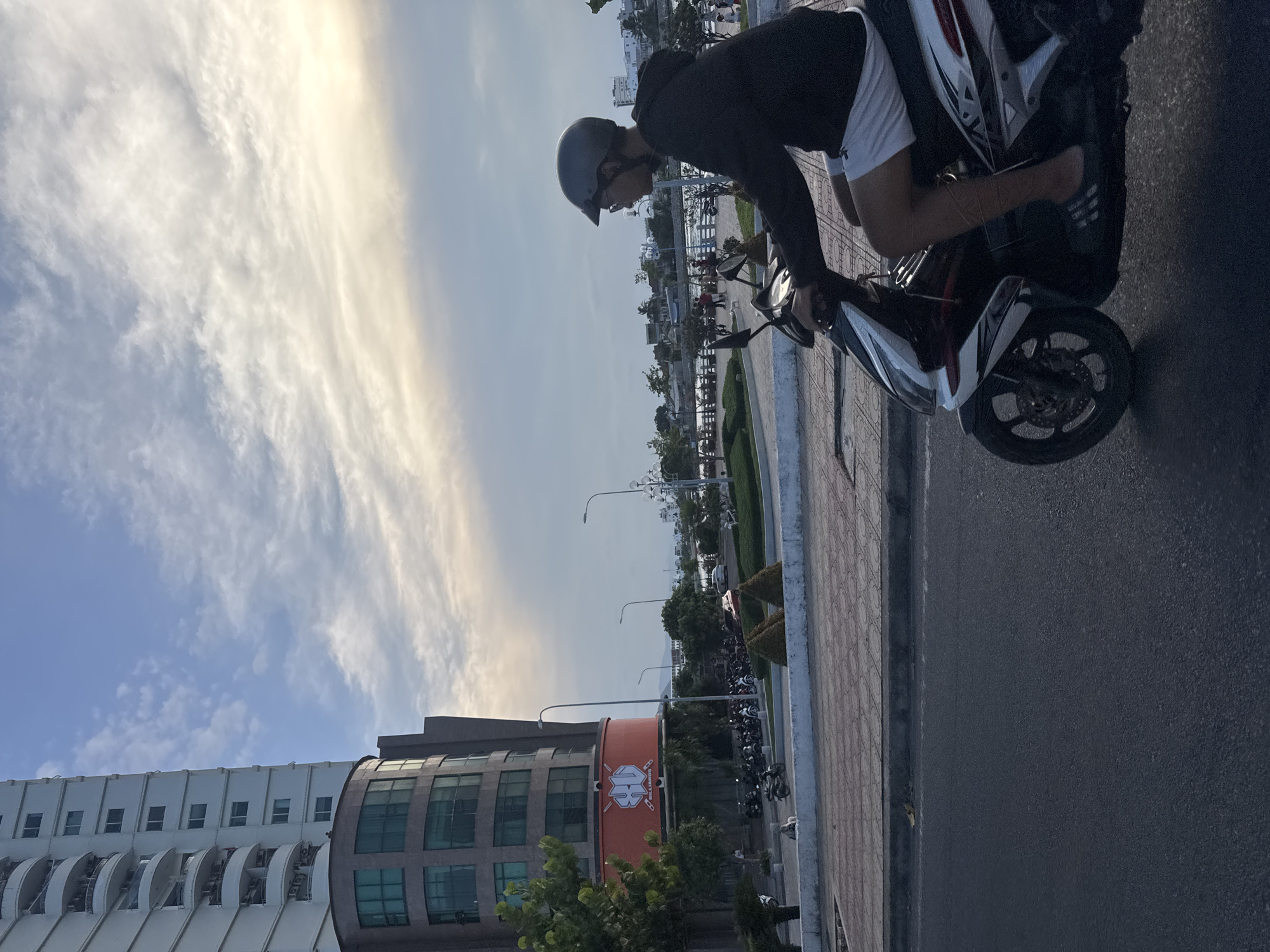 In Nha Trang, Vietnam, on a paved road next to a sidewalk, a young man wearing a dark hoodie, white shorts, and a grey helmet rides a white and black motorcycle. He is facing right, looking toward the horizon. In the background, a tall, white apartment building with balconies stands next to a curved, dark-glassed building with a large orange sign displaying the words "BU BILLIARDS." Beyond the buildings, a body of water, possibly the sea, is visible under a cloudy sky with hints of yellow light. Several motorcycles are parked in the distance.