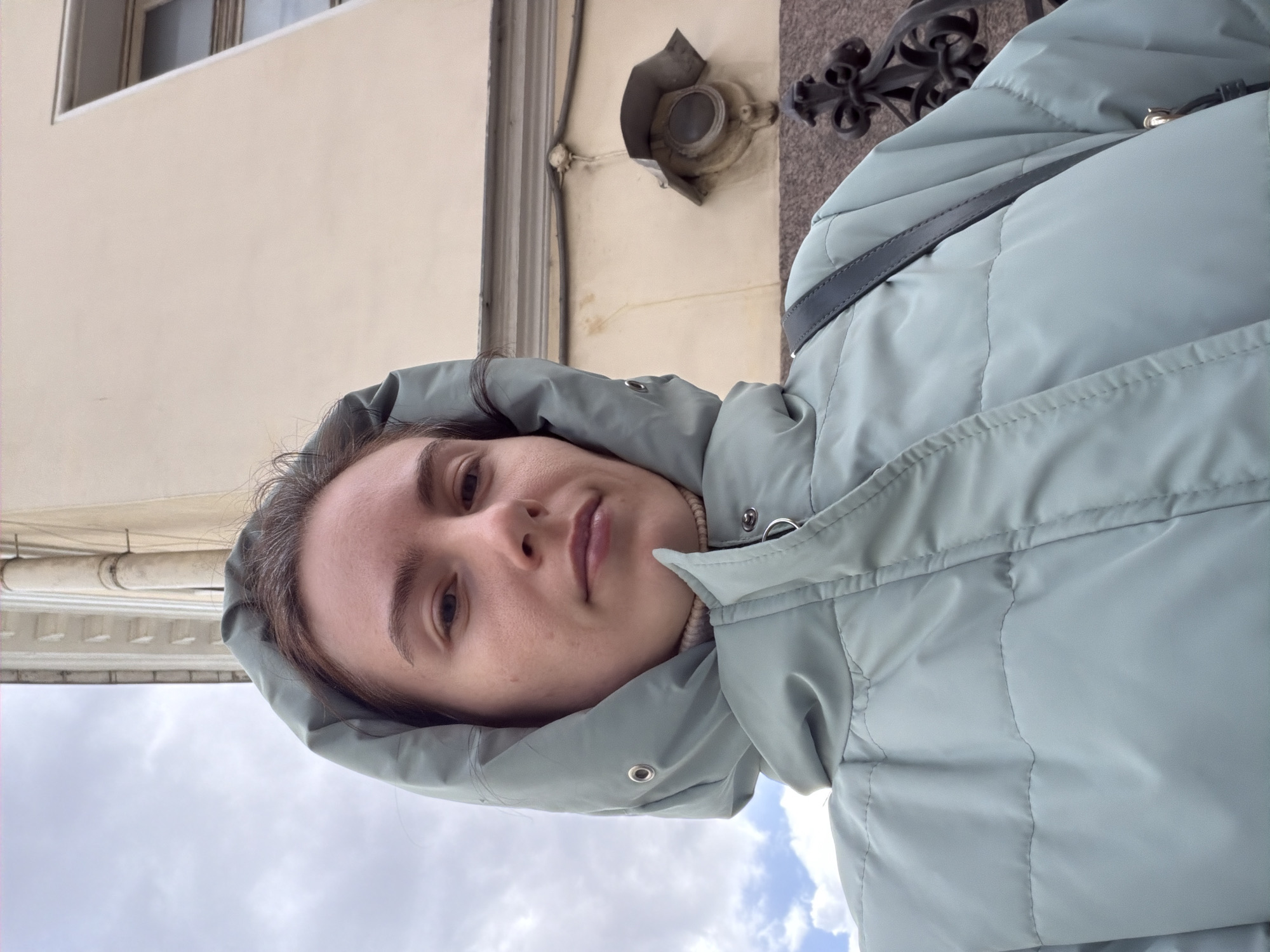 A woman in a light blue hooded puffer jacket is facing the camera. The background includes a light-colored building, likely stucco, with visible architectural details like window frames and cornices. A portion of an ornate black metal fence or railing is also visible. Based on the architecture and style, the location is likely Saint Petersburg, Russia. The photo appears to be taken from a slightly low angle.