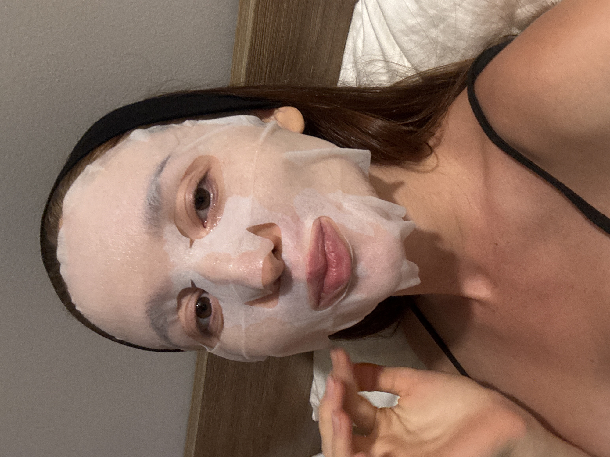 A woman is lying in bed with a sheet face mask on. She is wearing a black headband and a black tank top. Her hair is brown. She appears to be in a bedroom. The location is Choeng Thale, Thailand.