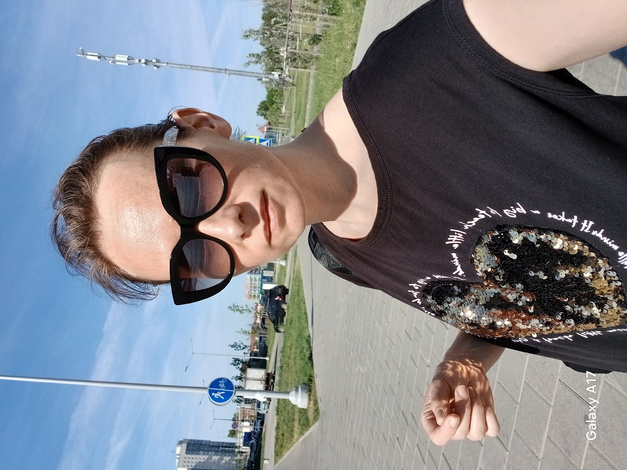 A middle-aged woman with short hair and large sunglasses is posing for a photo outdoors, likely in a public space. She is wearing a dark-colored t-shirt with a heart-shaped sequined design and white lettering around it. The photo is taken at a slightly low angle, and the camera is positioned to capture her face and upper torso.
The background indicates a city setting with a paved area, grassy patches, trees, street lights, and buildings in the distance. There's a blue road sign with a white bicycle and pedestrian symbol. A tall metal structure resembling a cellular tower or communication equipment stands prominently in the background. Based on visual clues such as architectural style and the signage, the location is potentially within Yekaterinburg, Russia. The overall lighting suggests a sunny day. The photo contains a watermark "Galaxy A17," implying the device used for taking the image. The woman's right hand is visible near the bottom of the image, positioned slightly clenched.