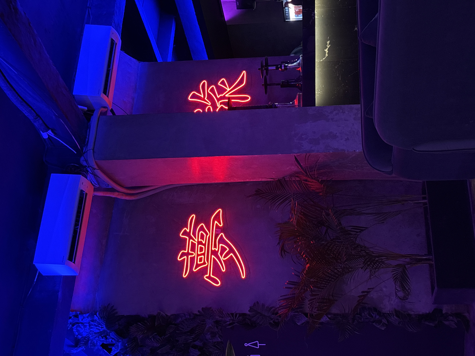 The scene depicts an indoor bar or lounge area in Kuta Selatan, Indonesia. Two walls are visible, each adorned with a neon sign in the style of Chinese or Japanese calligraphy. Above each sign, there is an air conditioning unit mounted. In the background, behind the top sign, there are bar fixtures, including taps. To the right of the lower sign, there are palm fronds and other tropical foliage, suggesting a themed or naturalistic decor. The lighting is predominantly blue and red, creating a modern and atmospheric ambiance. A chair is partially visible on the right side of the image.