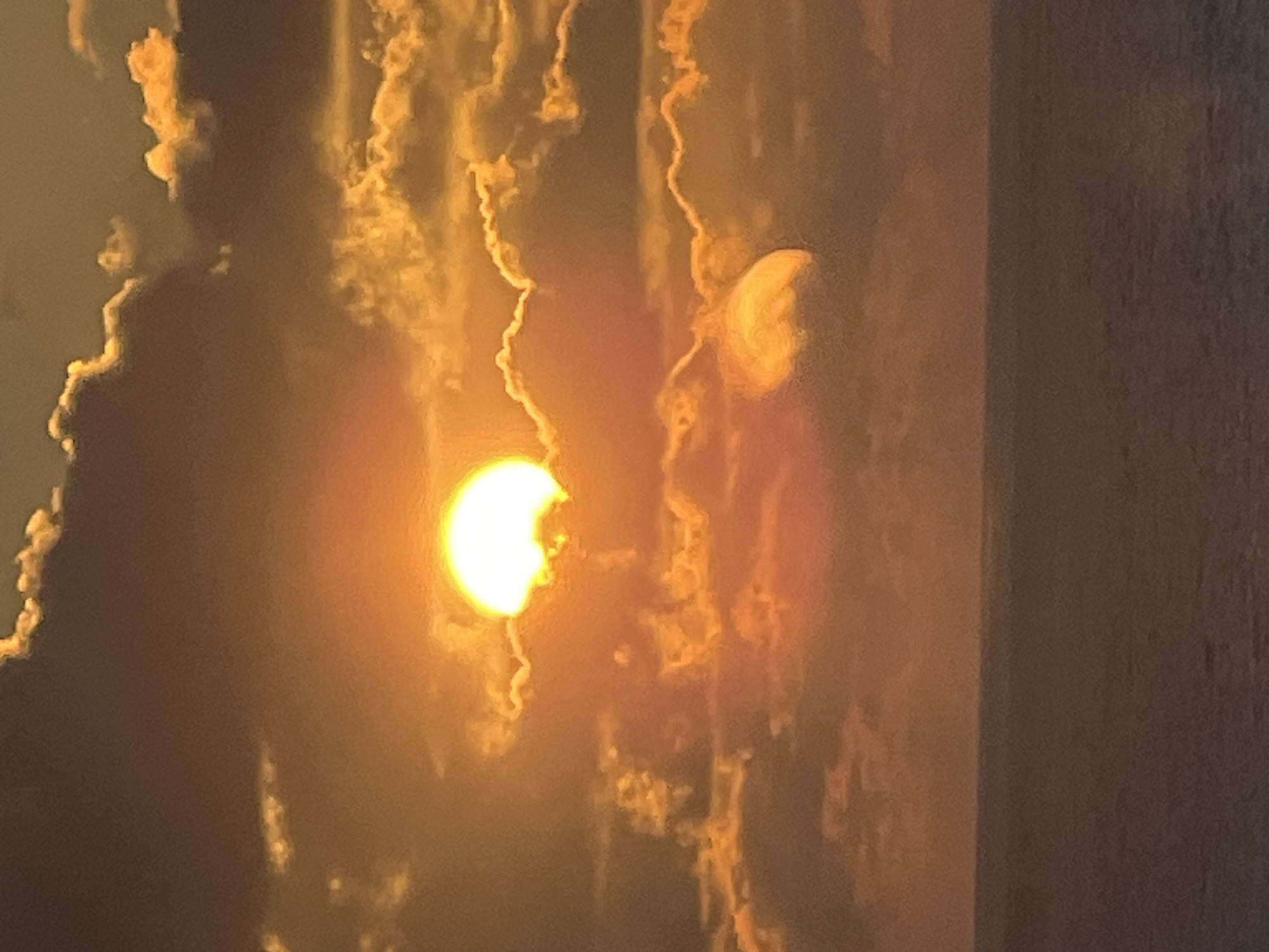 The image depicts a sunset over the ocean. The sun, a bright orb of light, is partially obscured by dark clouds, creating a dramatic contrast of light and shadow. The sky is filled with hues of orange, yellow, and brown, characteristic of a sunset. The ocean below is a muted, dark blue, with a subtle texture that suggests gentle waves. The horizon line is clearly defined, separating the sky from the water. The overall scene evokes a sense of calm and natural beauty.