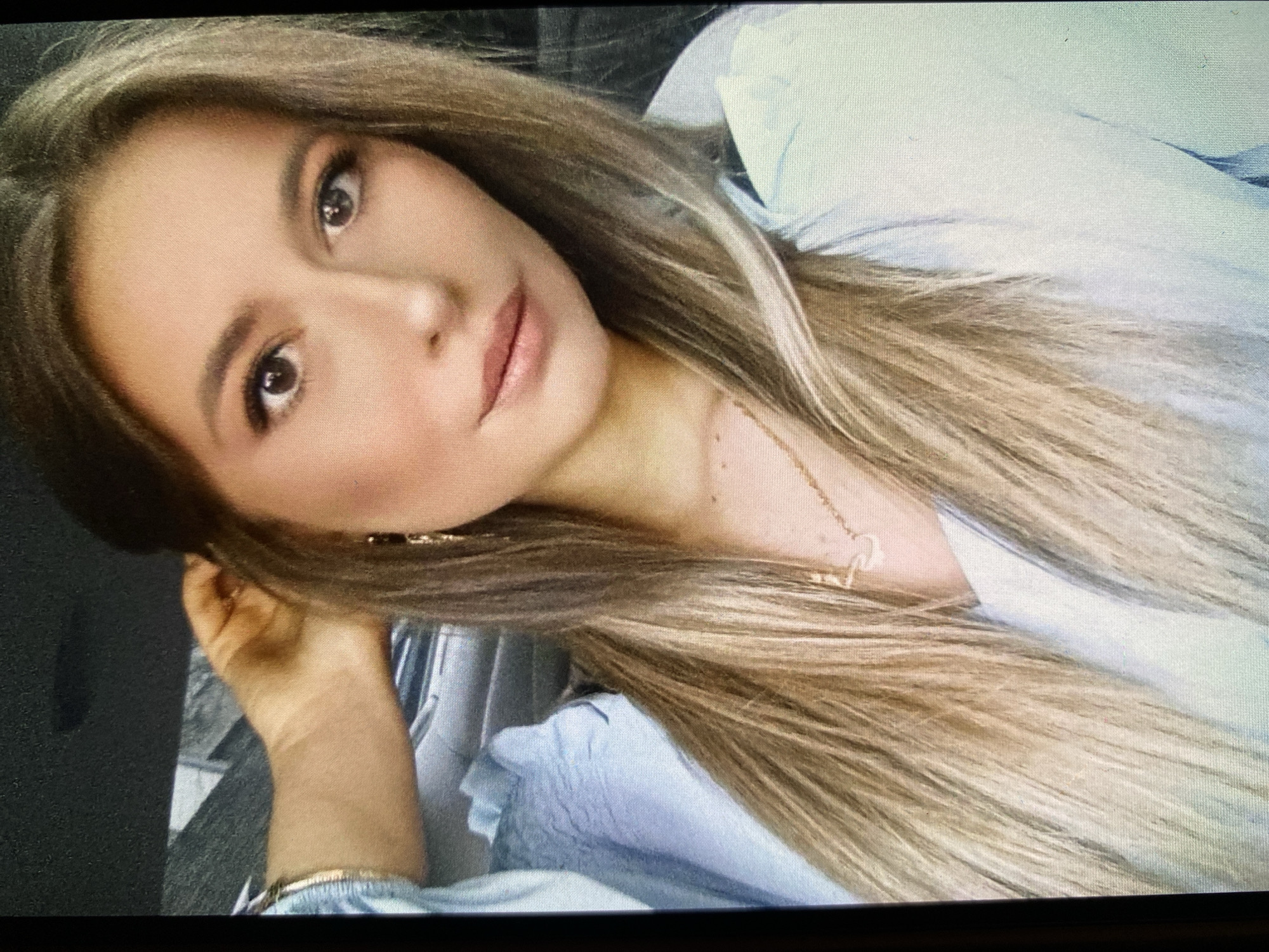 The image displays a close-up portrait of a young woman, taken in Bankya, Bulgaria. She is shown reclining or lying down, with her head tilted and looking directly at the viewer. Her long, straight hair, a blend of light brown and blonde highlights, frames her face and spreads across her shoulder.
The woman has fair skin and a calm, composed expression. Her prominent dark brown eyes are enhanced with mascara and eyeliner. Her eyebrows are well-defined, and her full lips are colored with a soft pink or nude lipstick. She wears a light blue, long-sleeved top, possibly a blouse or a light shirt, with some visible fabric gathers or ruffles on the sleeve near her left wrist.
Accessories include a delicate gold-colored chain necklace with a small pendant, and a thin gold-colored bracelet on her left wrist. Small gold-colored earrings are also partially visible. Her left hand is positioned near her head, possibly supporting it. The background is dark and out of focus, indicating an indoor setting. The lighting is soft and even, highlighting her features. There is no visible text in the image.