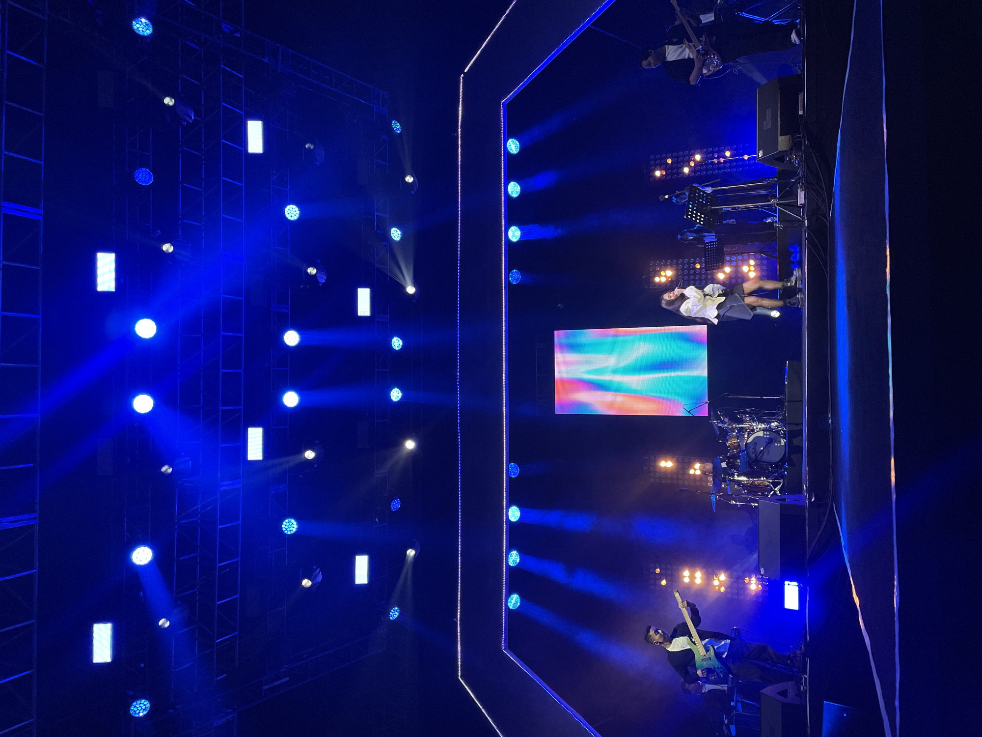 A stage is set in what appears to be a large venue in Salembaran, Indonesia, featuring a performance. Stage lighting is prominent with a blue color scheme, utilizing both spot and flood lighting techniques. The lighting rig is visible on the left. On the stage, there is a band performing, complete with a drummer, keyboardist, vocalist, and guitarist. A large screen is visible in the center-back of the stage displaying colorful graphics. The audience is not visible.
