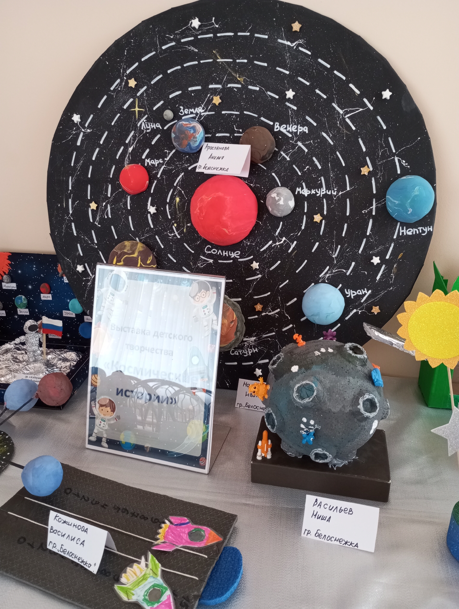 A display of children's space-themed art projects. A large circular black background, splattered with white paint to represent stars, serves as a backdrop for planets made of various materials like modeling clay. Each planet has a handwritten label in Russian, identifying it (e.g., "Sun," "Earth," "Mars"). One project features a diorama with a foil astronaut and the Russian flag on a small-scale planet. Another displays a textured gray planet with small astronaut figures attached. There are also name cards identifying the young artists: "Kozhinova Vasilisa," "Vasiliev Misha," and "Araslanova Amiliya," all from the town of "Belosnezhka." A framed sign reads "Exhibition of Children's Creativity 'Cosmic Stories'," indicating a school or local art exhibit in the city of Belosnezhka.