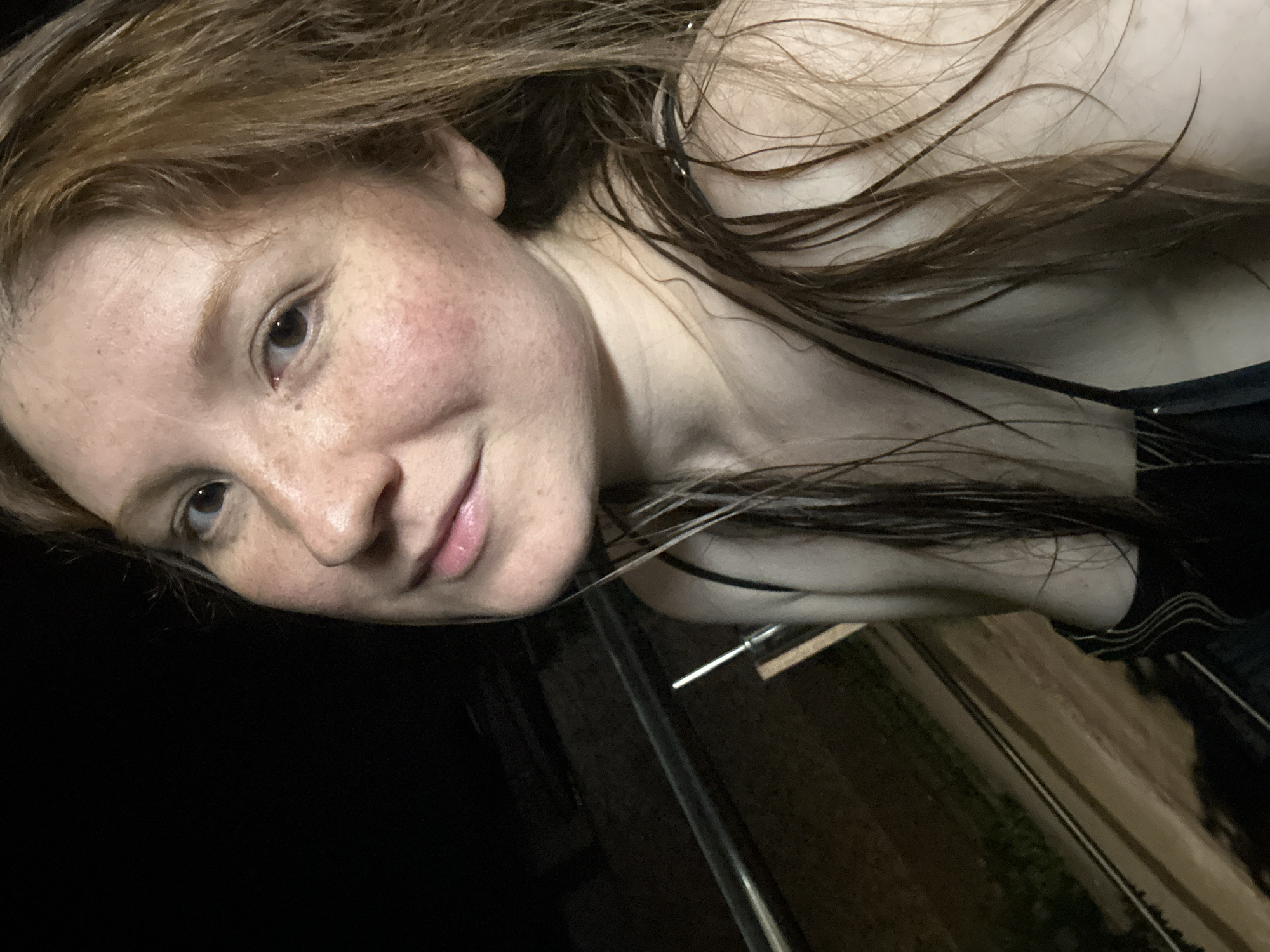 A selfie of a woman with freckles and wet red-brown hair, taken at night. The woman is fair-skinned with a light pink lip color. She is wearing a black spaghetti strap top. The background is dark, with hints of a reflective surface and some greenery. The exact location in Nizhny Novgorod, Russia, cannot be determined from this image.