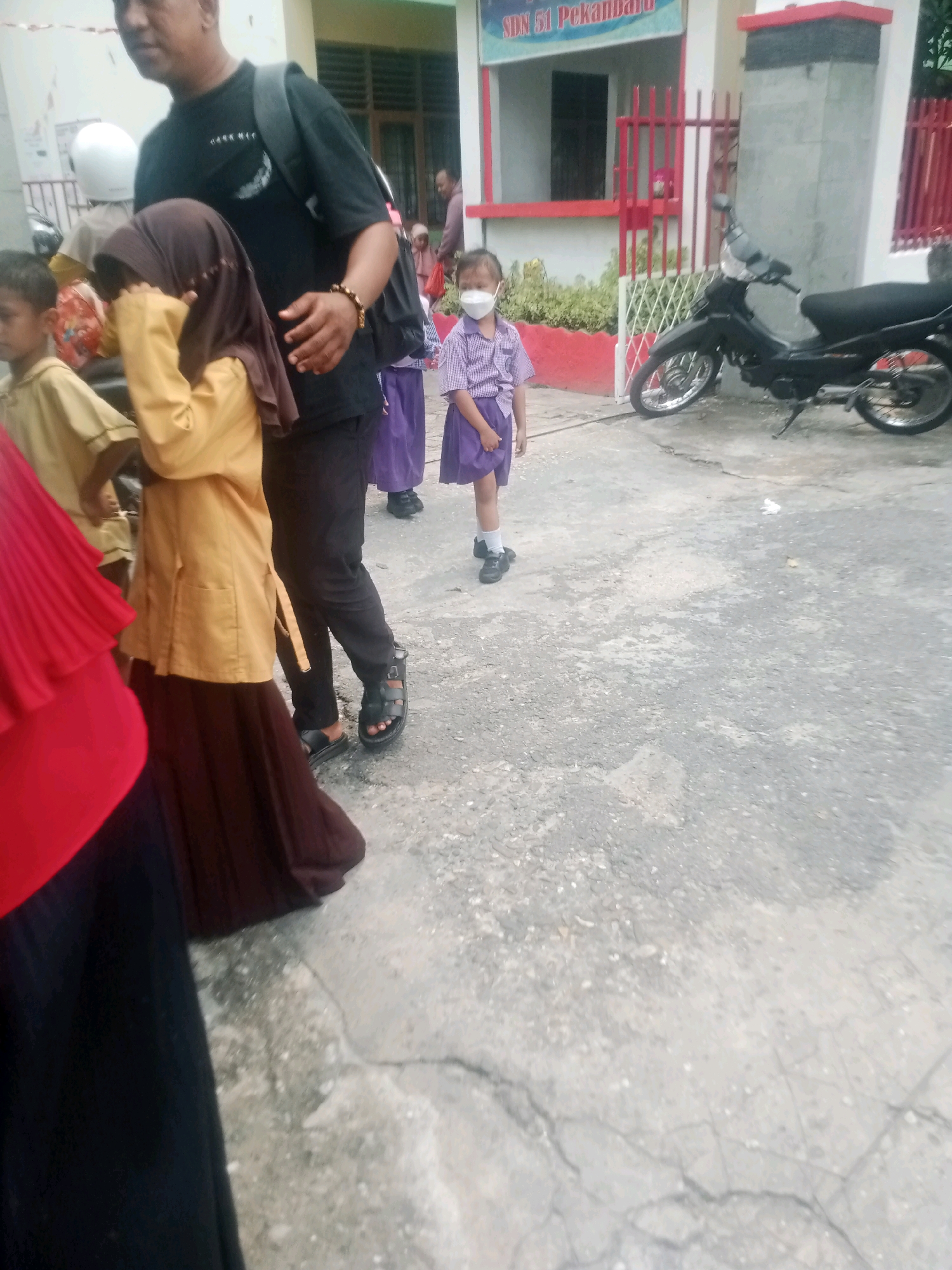 The image captures a scene outside a school in Pekanbaru, Indonesia, identified by a sign on the building reading "SDN 51 Pekanbaru." In the foreground, a person wearing a red hijab and black clothing is partially visible on the left, followed by a child in a mustard-colored outfit and a brown hijab, who is covering their face. To their right, a man in a black t-shirt and dark pants, carrying a backpack, stands next to the child. Further in the background, a young girl in a purple school uniform and a face mask is visible. A black motorcycle is parked on the right side of the frame. The environment appears to be an outdoor paved area, likely a school courtyard or walkway. The overall scene suggests a moment of children and adults gathered outside the school, possibly during arrival or dismissal.