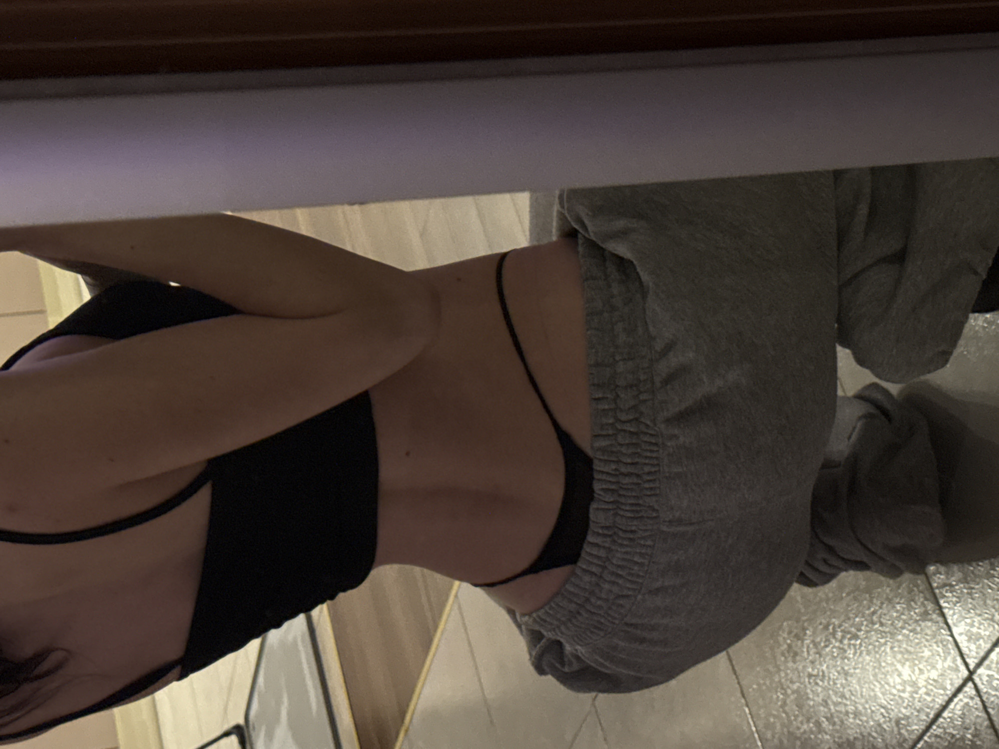 The image shows a close-up, rear view of a person squatting in what appears to be an indoor setting. The person is wearing a black crop top, grey sweatpants, and a black thong. The background consists of a tiled floor and a reflective surface, possibly a mirror or glass, showing a faint reflection of the person's arm and shoulder. The overall lighting is dim, with some light sources creating reflections on the floor. There are no clear indicators of a specific city or outdoor environment.
