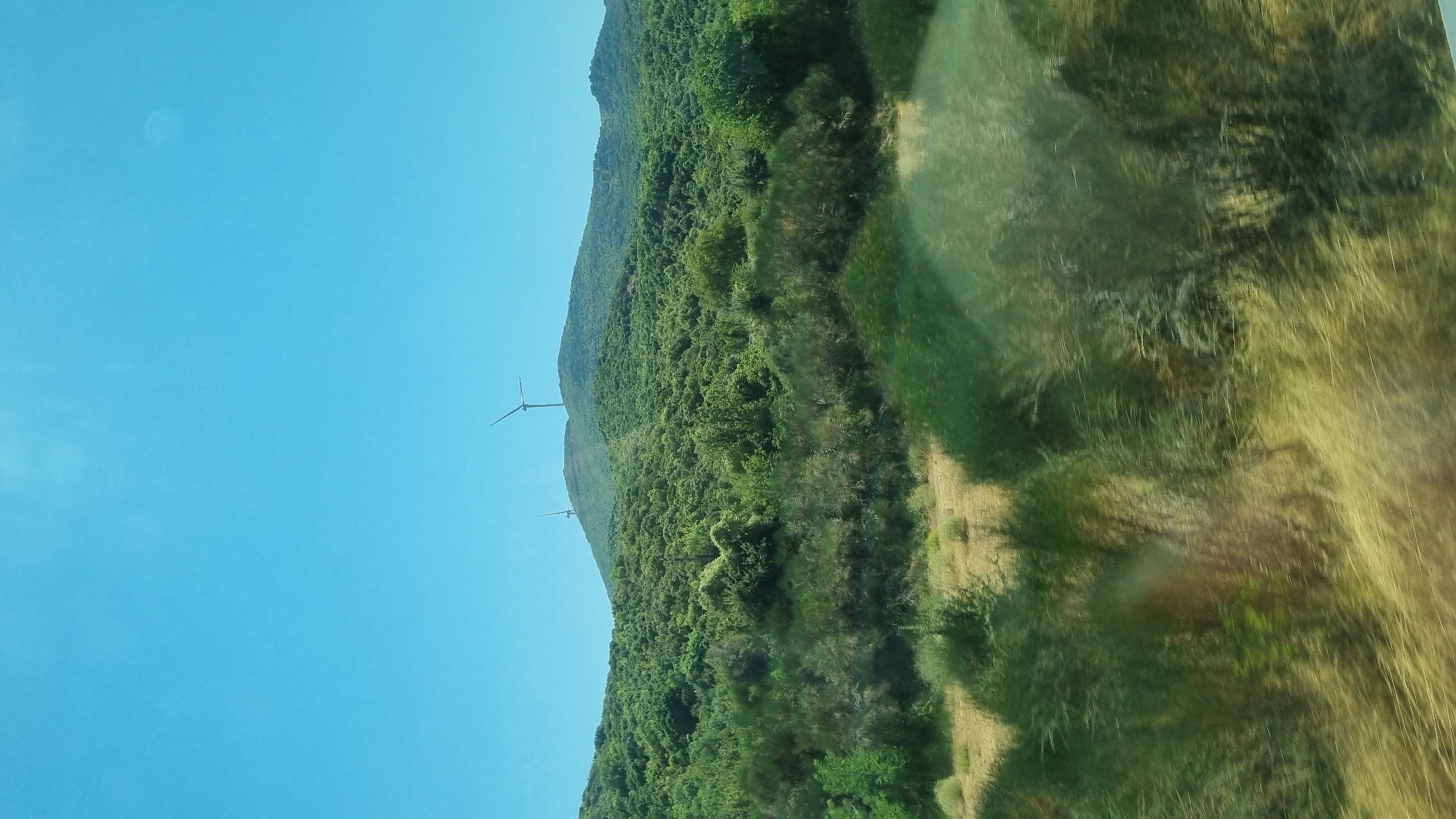 This is a landscape photograph taken from a moving vehicle, looking towards a hillside covered in dense green vegetation. Two wind turbines are visible on the crest of the hill against a clear blue sky. The foreground consists of blurred vegetation, suggesting a motion effect. The overall scene depicts a rural or semi-rural environment in Greece, possibly in the municipal unit of Promachonas.