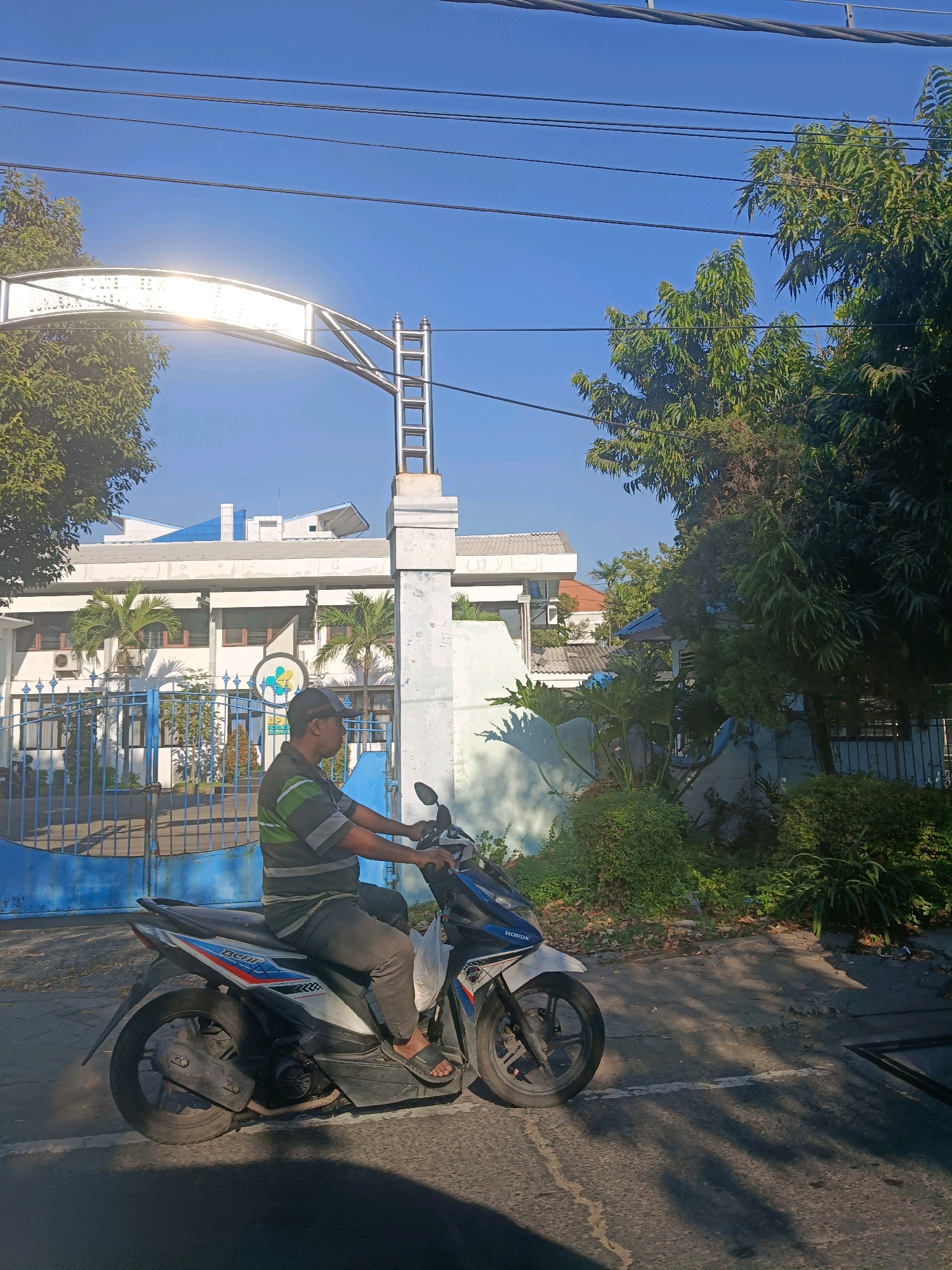 A man wearing a dark grey and green striped shirt, dark pants, and black sandals is riding a white and blue Honda Beat motorcycle. He is wearing a black baseball cap. The motorcycle is moving on a paved road. The man is looking forward. In the background, there is a large white building with blue accents and a blue metal gate. A large arched sign is visible above the gate. Trees and other foliage are present around the building and gate. The sky is clear and blue. The image appears to have been taken in Surabaya, Indonesia, likely on a sunny day.