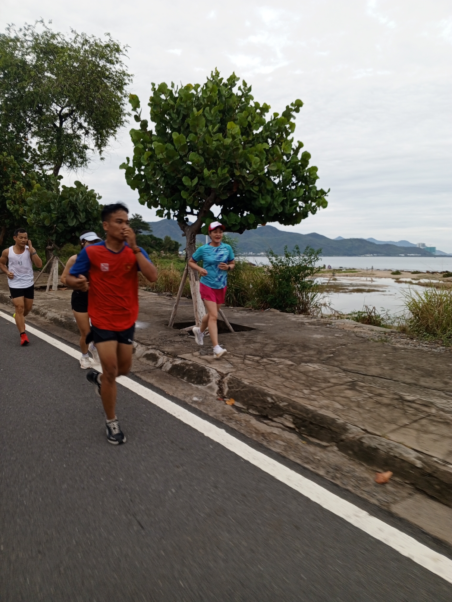 The image captures a scene in Bac Nha Trang Ward, Vietnam, featuring four people jogging along a road next to a body of water. The runner closest to the camera wears a red shirt with blue sleeves, dark shorts, and running shoes. Behind him to the left, another runner is wearing a black tank top and shorts. To the left of that runner, a man runs in a white tank top and black shorts with red running shoes. Slightly ahead of the first runner, to the right, a woman wearing a light blue shirt and pink shorts jogs. The group runs along a road marked with a white line. To the right of the road is a concrete sidewalk bordered by vegetation. The body of water is visible beyond the sidewalk, with mountains in the distance. Trees frame the scene on either side. The sky is overcast.