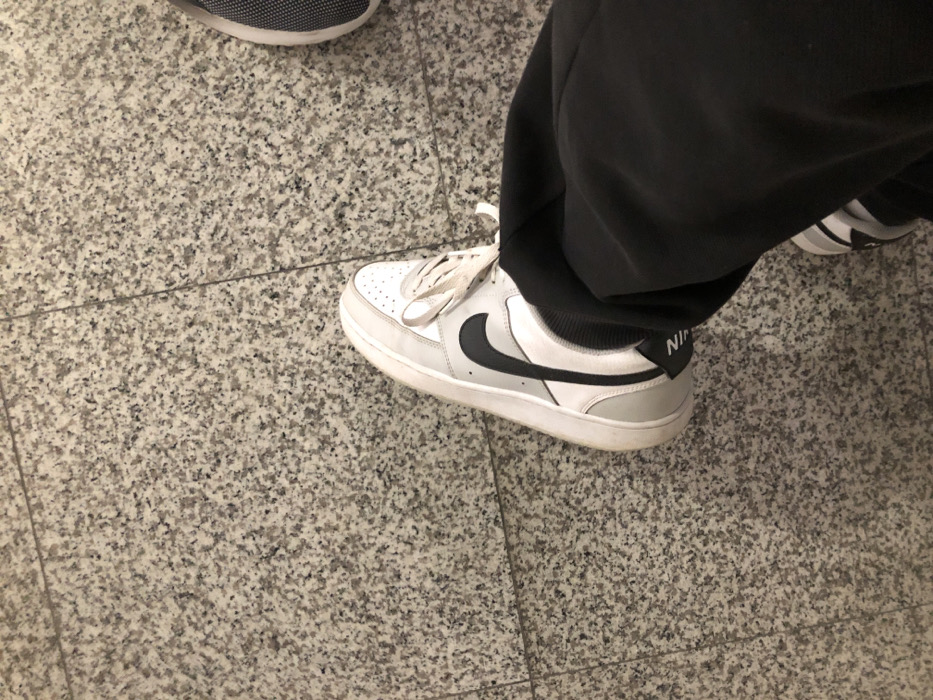 The image captures a ground-level view of two people's feet on a speckled light grey and black tiled floor, likely indoors. The location is Moschato, Greece, though no specific visual cues within the image confirm this.
In the foreground and central to the image is a person's right foot, wearing a pair of white and grey Nike sneakers and dark grey or black sweatpants. The sneaker features a white perforated toe box, light grey side panels, and a prominent black Nike swoosh. The laces are white and appear somewhat loose or untied. The shoe's white midsole shows minor signs of wear. On the grey heel tab, the word "NIKE" is visibly printed in white capital letters. The black sweatpants are loose-fitting, covering the ankle and partially resting on the top of the shoe.
In the upper left corner, the toe and side of another person's shoe are partially visible. This shoe appears to be a dark grey or black sneaker with a textured or mesh upper and a white sole. The activity depicted is simply people standing or walking on a tiled floor. No specific event or time of day is discernible, and no emotions can be interpreted as faces are not visible.