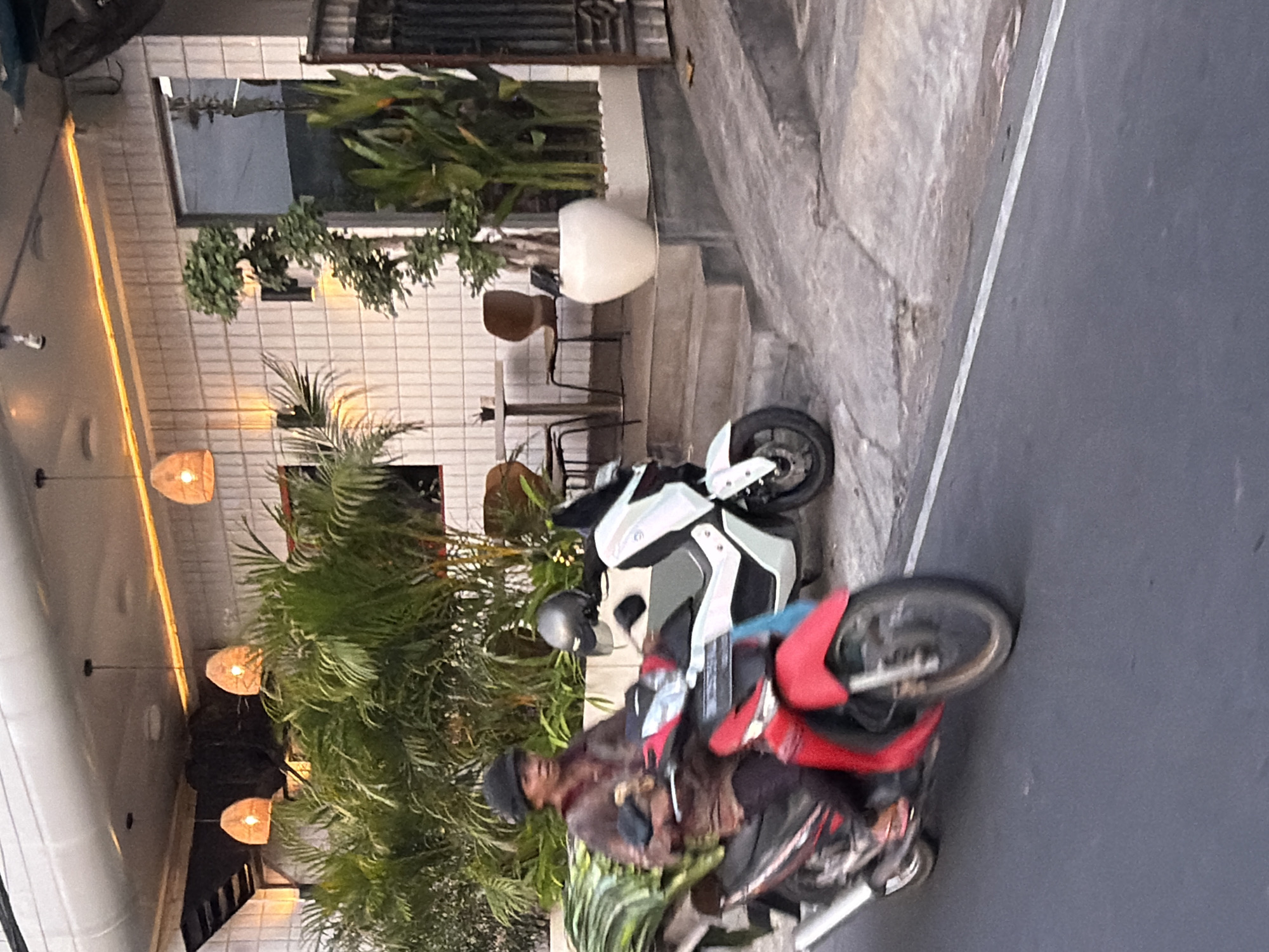 The image captures a street scene in Mengwi, Indonesia, likely during the day. Two motorcycles are prominently featured. The first, closer to the foreground, is a red and black scooter with a rider wearing a helmet and a brown jacket. The rider appears to be holding something in their hands. Behind this scooter is a white and gray sportbike, parked or stationary.
The background reveals a building with white tiled walls and large windows. Several potted plants, including lush green foliage, are visible, suggesting an outdoor seating area or garden associated with the building. Three woven pendant lights hang from the ceiling, illuminating the area with a warm glow, indicating it might be an eatery or a commercial establishment. The ground is paved with concrete and asphalt. The lighting suggests it is daytime, possibly late afternoon given the warm tones of the lights. There is no visible text in the image.