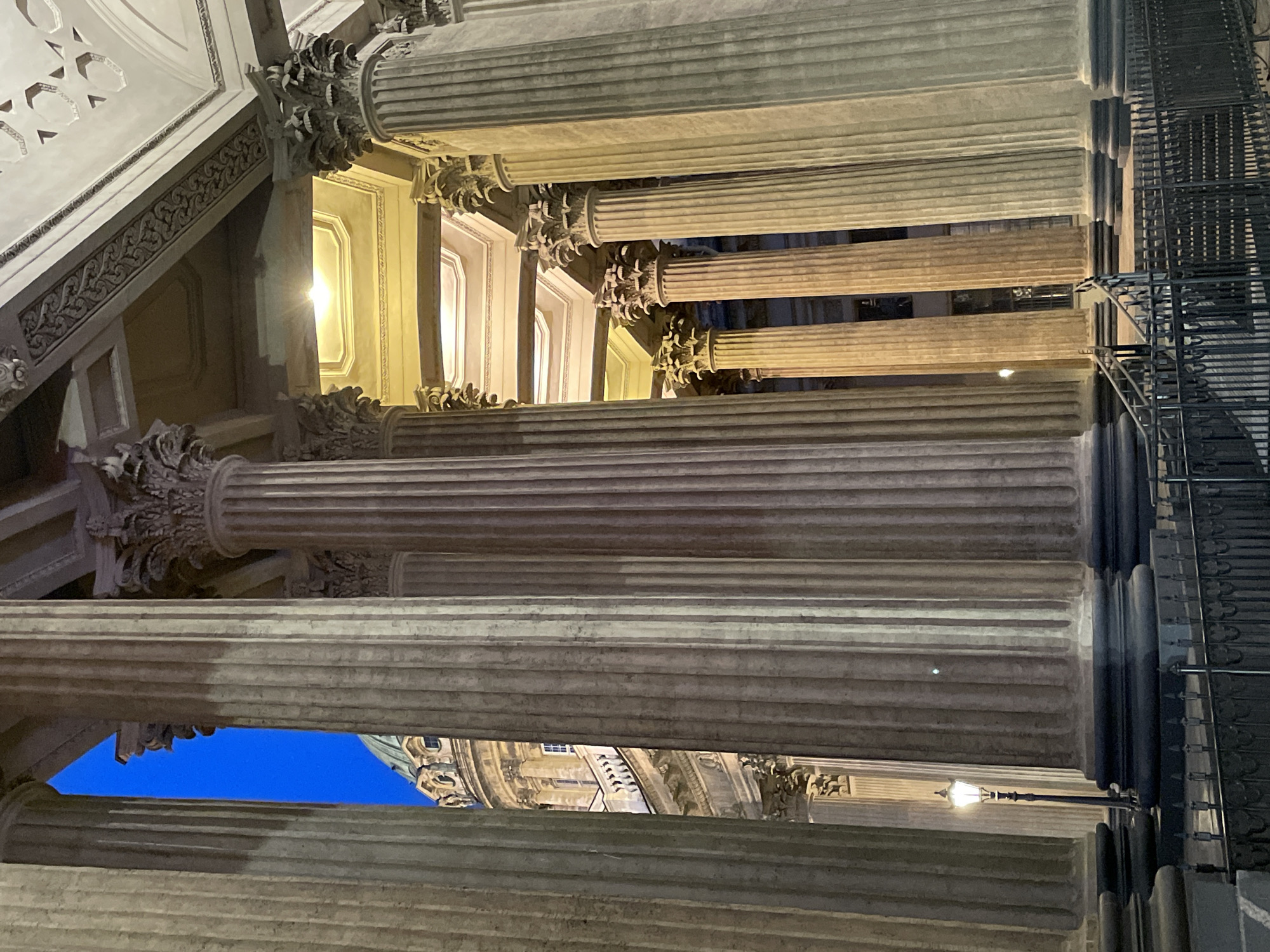 This image depicts the colonnade of the Kazan Cathedral in Saint Petersburg, Russia, observed during evening or night hours. The scene is dominated by numerous large, fluted Corinthian columns, constructed from a light-colored stone, likely granite or marble. These columns exhibit varying hues from cool gray to warm amber and yellow due to diverse artificial light sources illuminating them and the surrounding architecture.
Through gaps in the colonnade, a narrow vertical section of deep blue sky is visible on the left. To the right of this, and seen more extensively through other column intervals, is the upper portion of a large, green-domed structure, identifiable as part of the Kazan Cathedral's main rotunda. This dome features multiple windows, balustrades, and detailed architectural ornamentation, illuminated with a warm, yellowish light.
The ceiling structure above the colonnade is visible, showcasing decorative entablature and coffered sections. These areas are illuminated by recessed artificial lights, casting a warm glow and revealing intricate carvings and geometric patterns, including star-like motifs.
At the base of the scene, a dark metal fence, likely wrought iron, runs horizontally across the foreground. To the left, a classic black streetlamp with an illuminated yellow globe stands between the fence and the initial row of columns. On the right, a set of stone stairs with a metal handrail leads upwards behind the fence, towards the elevated base of the colonnade. No individuals are present in the image.