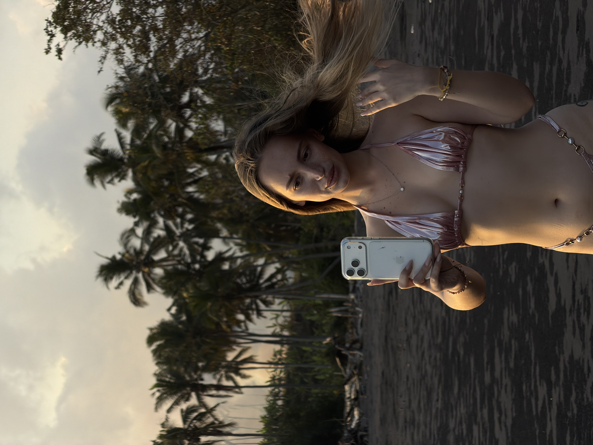 A blonde woman wearing a pink metallic bikini takes a selfie with a smartphone while standing on a dark sand beach. She is holding the phone in her right hand and her left hand is near her hair, which is being blown by the wind. The background features a line of palm trees and lush vegetation against a cloudy sky at sunset. The scene is likely located in Selemadeg Timur, Indonesia, a region known for its beaches and tropical landscapes.