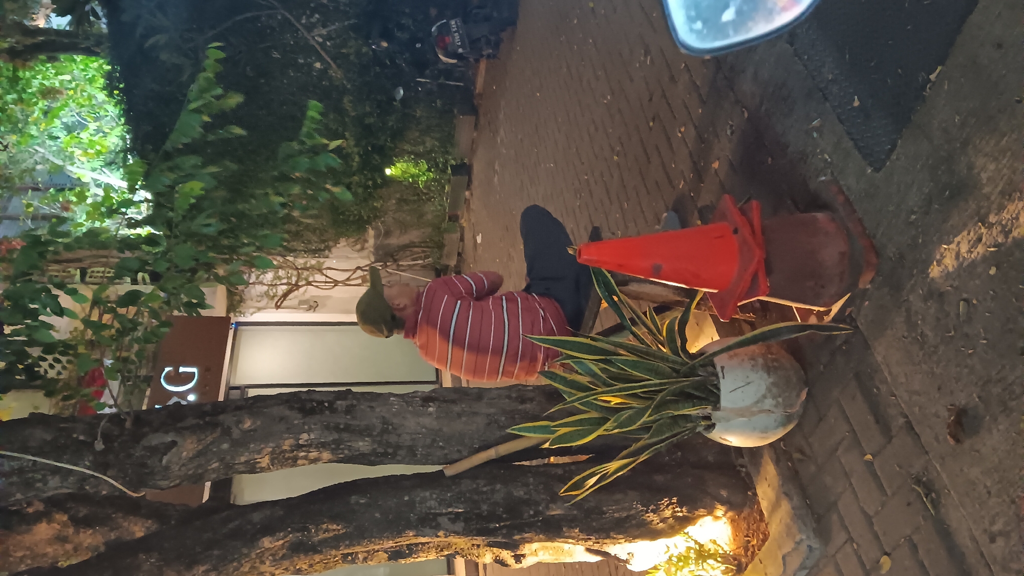 The image shows an outdoor scene at night on a brick-paved walkway in South Jakarta, Indonesia. A man wearing a green cap and a red and white striped shirt is sitting on a bench next to a tree. In front of the bench are a potted snake plant and an orange traffic cone. In the background, there is a building with a sign that reads "G" and a motorcycle parked in front of a hedge. The lighting suggests artificial illumination from streetlights or building lights.