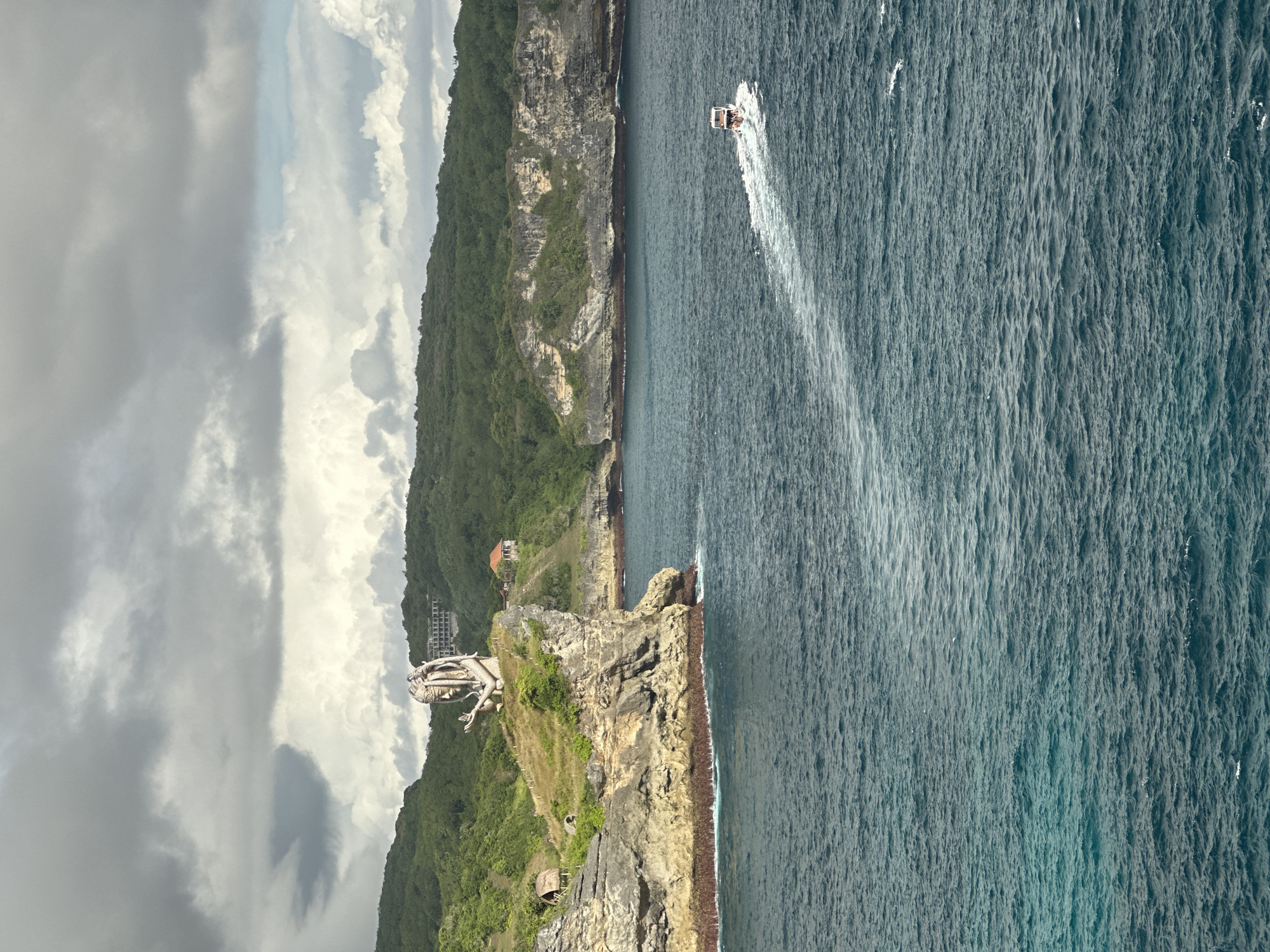 The image captures a coastal landscape in Nusa Penida, Indonesia. Dominating the left side is a rugged coastline densely covered in green vegetation, sloping down to a rocky cliff face. On the cliff, there is a large structure that is likely a building or a statue. Adjacent to the cliff is the vast expanse of the ocean. A single boat is visible, leaving a wake in the water. The sky overhead is filled with clouds, suggesting overcast weather.