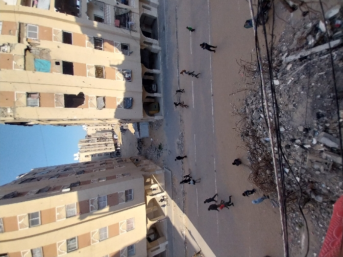 The image displays a damaged urban environment in Khan Yunis, Palestinian Territory, under a clear blue sky. Two multi-story residential buildings, constructed of beige concrete with brown accents, frame a paved street. The building on the right exhibits severe structural damage, including large sections of walls missing and multiple shattered or obscured windows, revealing interiors and a blue tarp. The building on the left also shows signs of damage and boarded windows. The ground floors of both buildings feature arched pedestrian access points; a yellow vehicle and a dark vehicle are parked beneath the arches of the right building.
Approximately 15-20 individuals are present on the street and adjacent areas. A prominent group of about ten people is seen walking or jogging across the paved street from various directions. Other individuals are distributed on the unpaved ground; one person is seated on the ground near a rubble pile in the foreground, and two smaller figures are observed near a tricycle or cart by the base of the left building.
The immediate foreground consists of a large pile of concrete debris, twisted rebar, and rubble. Overhead, black cables are suspended across the lower frame. The paved street appears dusty or dirty, and unpaved verges flank its sides. Strong sunlight creates distinct shadows, indicating a bright daytime setting. The scene reflects significant destruction and an active presence of civilians navigating the altered landscape.