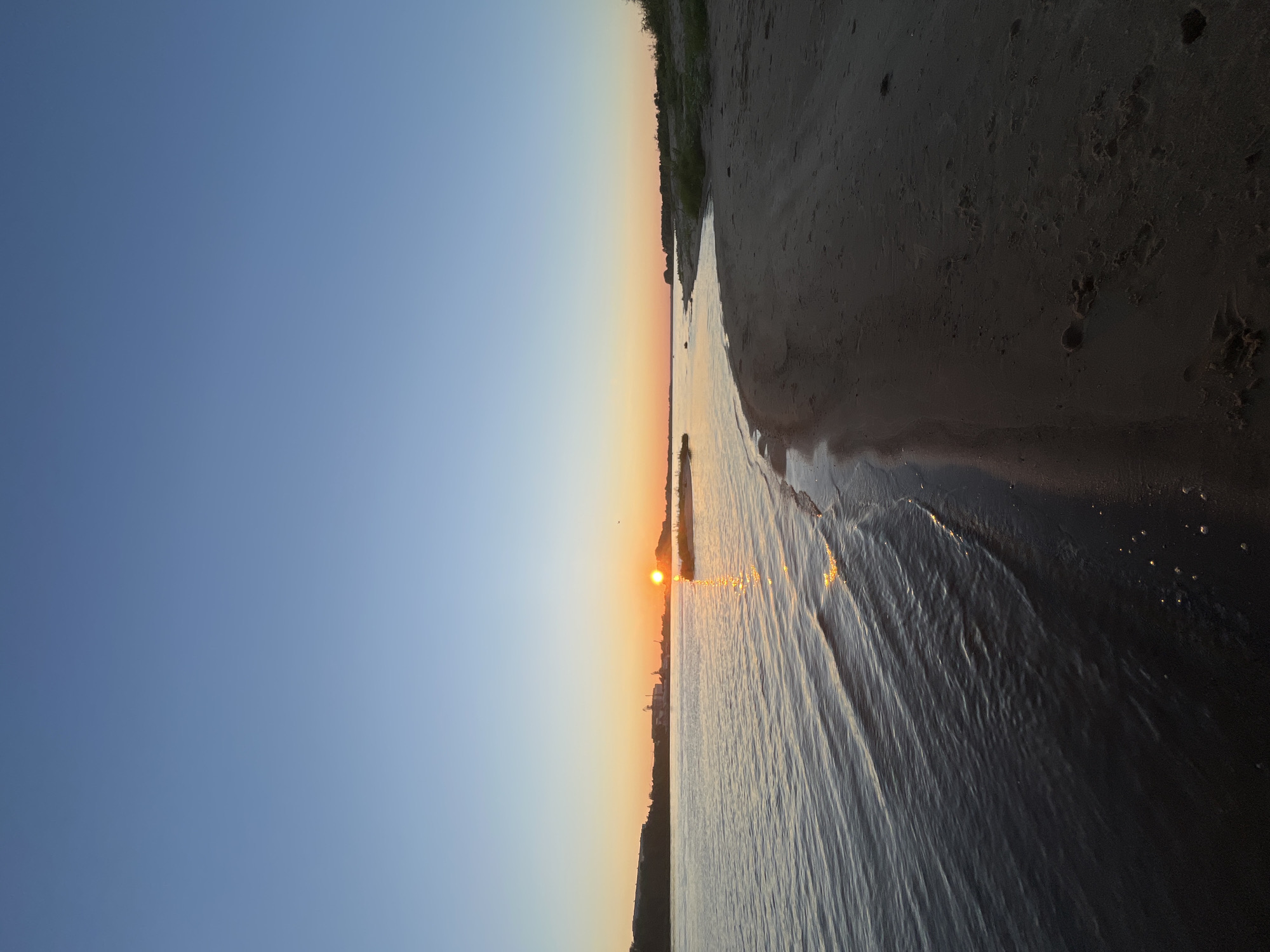 The image captures a sunset over a wide body of water, likely the Volga River, with a sandy shoreline in the foreground. The sky is a clear gradient of light blue to soft orange near the horizon. The sun, a bright orb, is setting behind a distant landmass with industrial structures visible. The water reflects the sunset's colors, creating shimmering streaks on its surface. The sandy bank is dark and appears to have some footprints. The environment suggests a natural setting on the outskirts of Nizhny Novgorod, Russia, possibly near a port or industrial area given the distant structures.