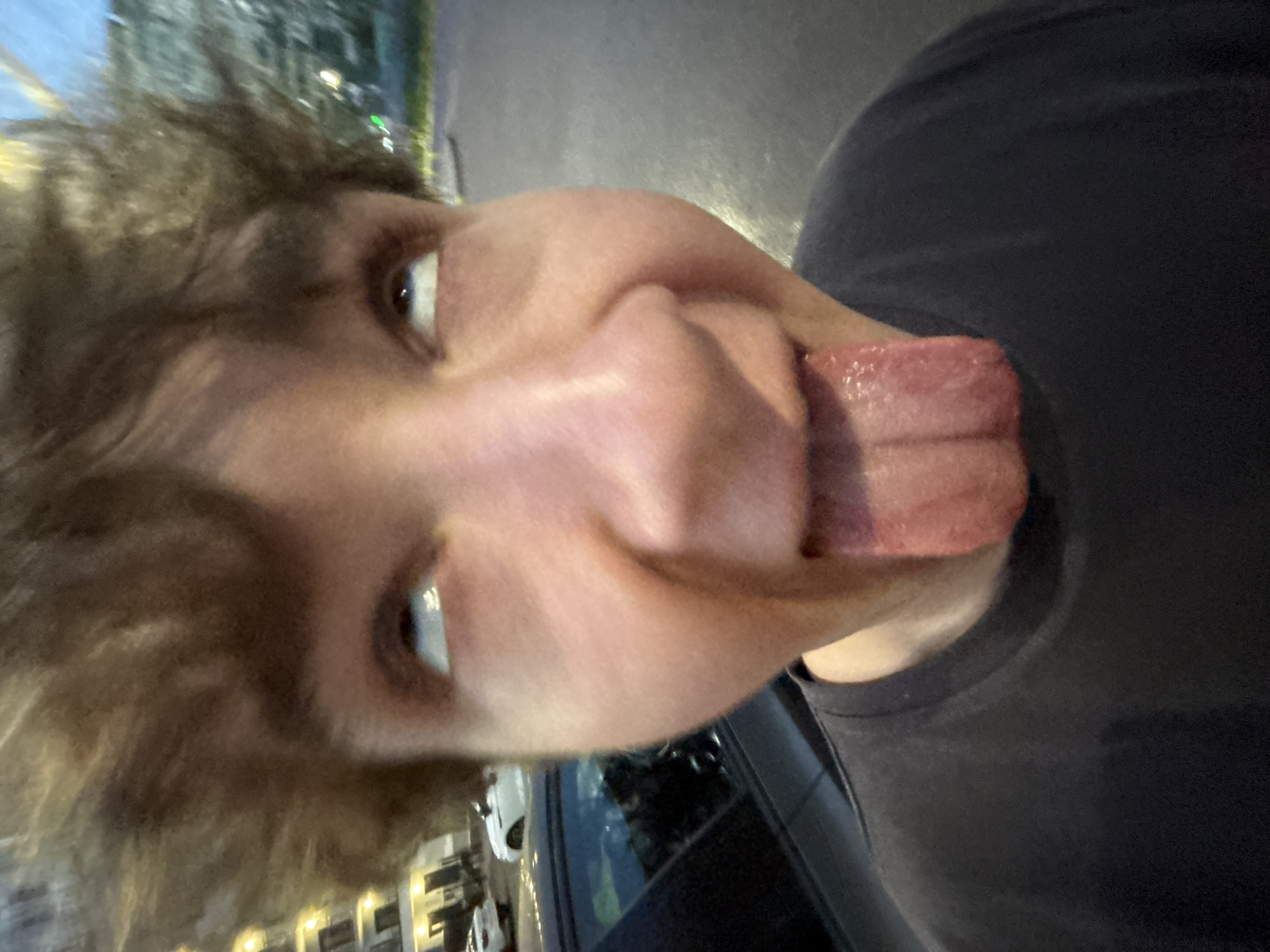 The image shows a close-up, distorted, and rotated view of a person's face. The subject is sticking out their tongue, which is very wide and appears to have some moisture on it. The person's eyes are visible and open, looking slightly upwards. Their hair is messy and appears light brown or blonde. They are wearing a dark t-shirt. The background is blurred, but it suggests an outdoor setting at night or in low light. There are some out-of-focus lights and reflections visible, possibly from buildings or streetlights, and a car is partially visible in the lower right portion of the image. The overall impression is that of a playful or goofy selfie taken at night. There is no visible text in the image.