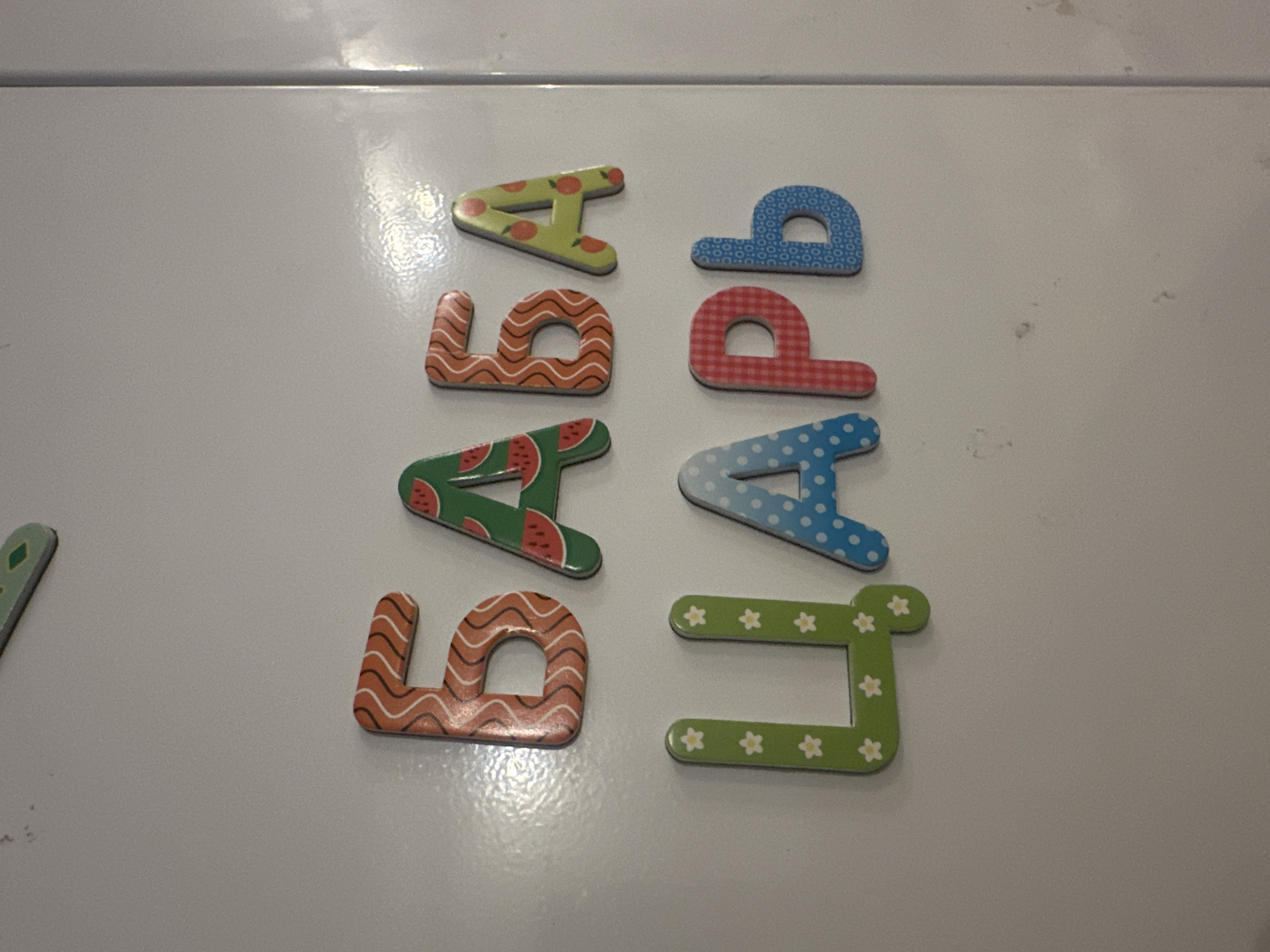 The image shows a collection of magnetic letters arranged in two vertical columns on a white surface. The letters are in pairs, one green and one orange, or red, or blue. The objects are patterned with various designs such as watermelons, flowers, polka dots, zigzags, and gingham. The letters are on what appears to be a white refrigerator or similar appliance. The location of Krasnaya Polyana, Russia is not apparent from the image.