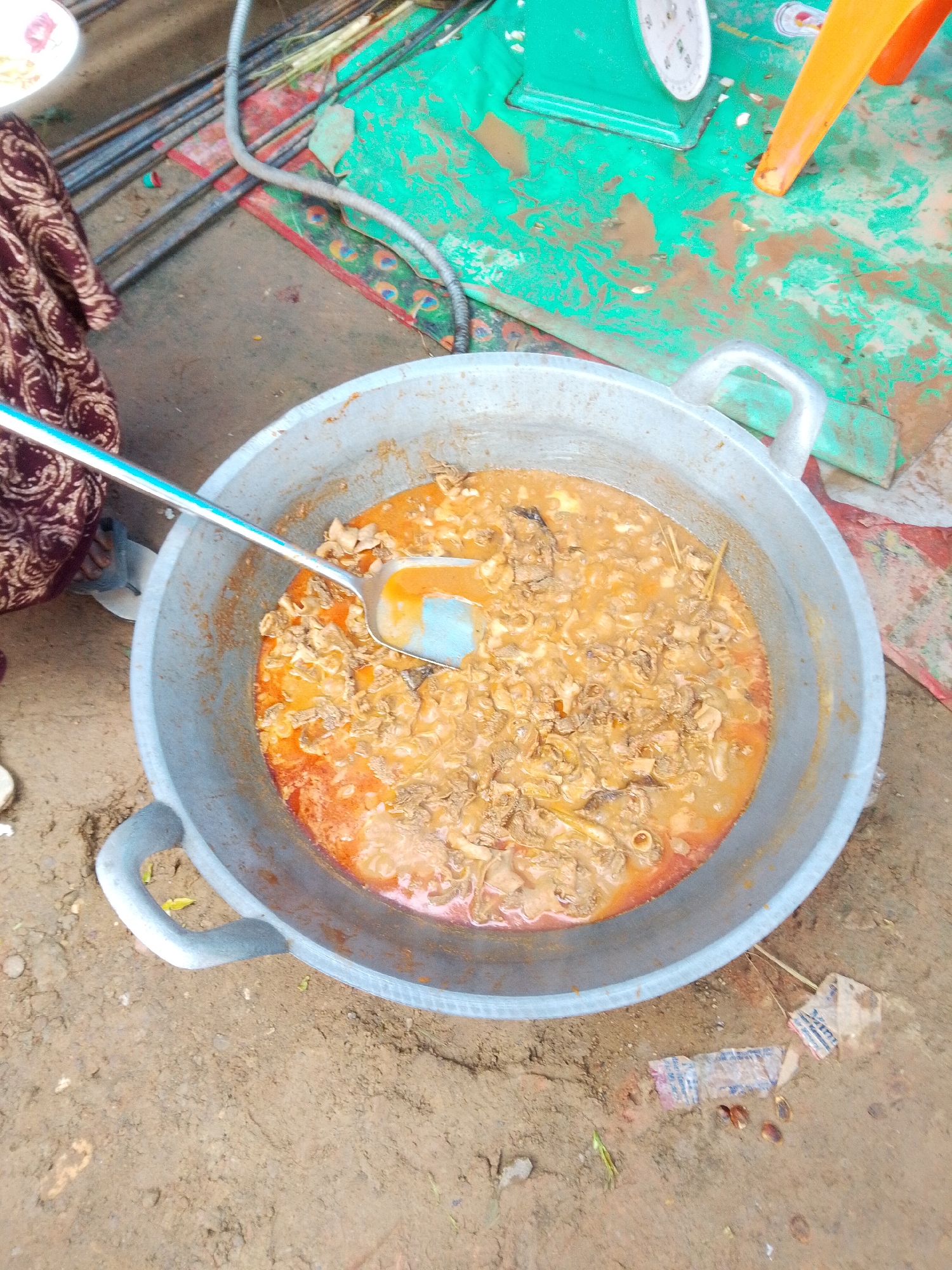 A close-up, overhead view shows a large, metal cooking pot filled with a savory stew. The stew is a rich, reddish-brown color and appears to contain chunks of meat and possibly vegetables. A large, silver ladle with a blue handle is partially submerged in the stew, suggesting it is being stirred or served. The pot rests on a dirt or sandy ground, with some scattered debris and small green leaves visible around it. In the background, a bright green tarp or plastic sheeting is visible, along with some metallic rods. A portion of a person's maroon and white patterned clothing is also visible on the left side of the frame. The scene appears to be an outdoor cooking or food preparation area.