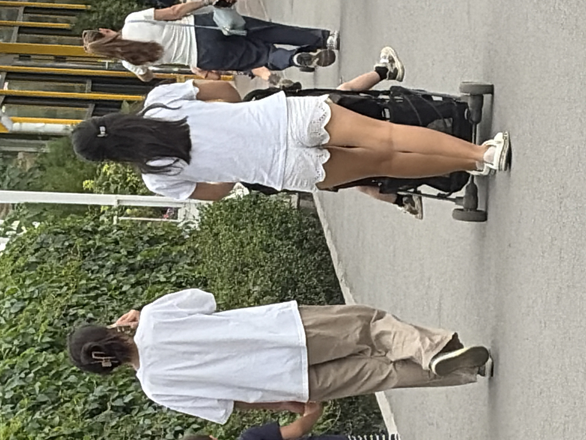Two women and a child are walking on a paved path next to a dense green hedge. The woman on the left, seen from behind, wears a white t-shirt and tan pants. She has dark hair pulled up with a clip and is holding a phone to her ear. To her right, a woman also seen from behind, wears a white t-shirt and white lace shorts. She is pushing a stroller with a child. The child's legs and feet are visible, wearing black socks and patterned sneakers. Another woman in a white t-shirt and dark pants is walking in the background to the right, carrying a light blue bag. The environment appears to be an outdoor public space with a building with yellow vertical supports visible in the far background.