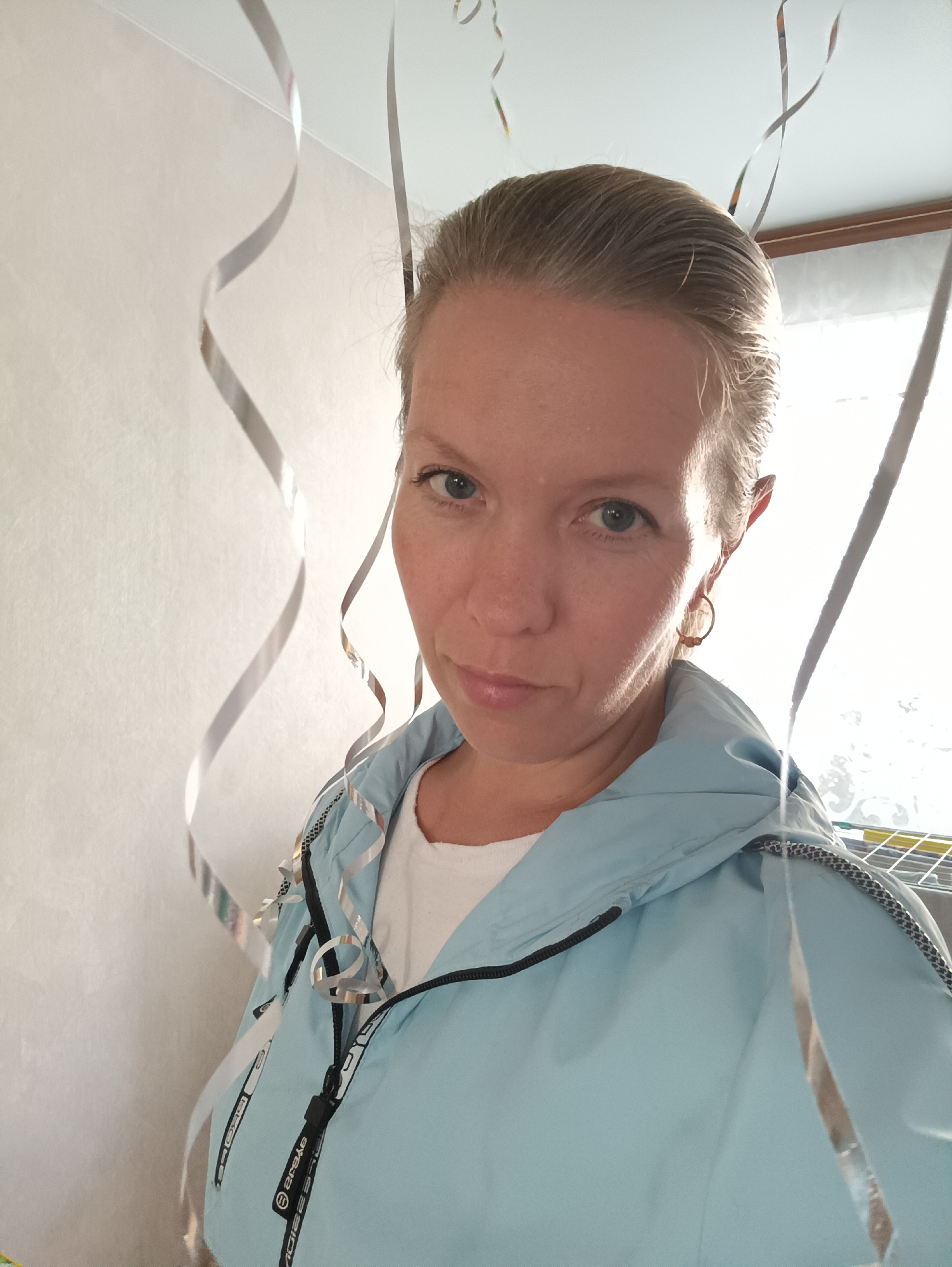 A woman is depicted in a close-up, half-body portrait taken indoors in Chaikovsky, Russia. She has light-colored hair pulled back from her face, light blue eyes, and a neutral, slightly contemplative expression as she looks directly at the camera. She is wearing a light blue jacket with black trim and zippers over a white t-shirt. A small, gold-toned hoop earring is visible on her right ear.
The background features light-colored walls, with several shiny silver streamers or ribbons hanging around the woman, suggesting a festive or celebratory occasion. Bright daylight streams in from a window on the right side of the frame, indicating the photo was taken during the day. Through the window, faint outlines of what appears to be a clothes drying rack can be seen. Visible text on the zipper pull of her jacket, and also on a black band along the jacket's trim, clearly reads "arcys".