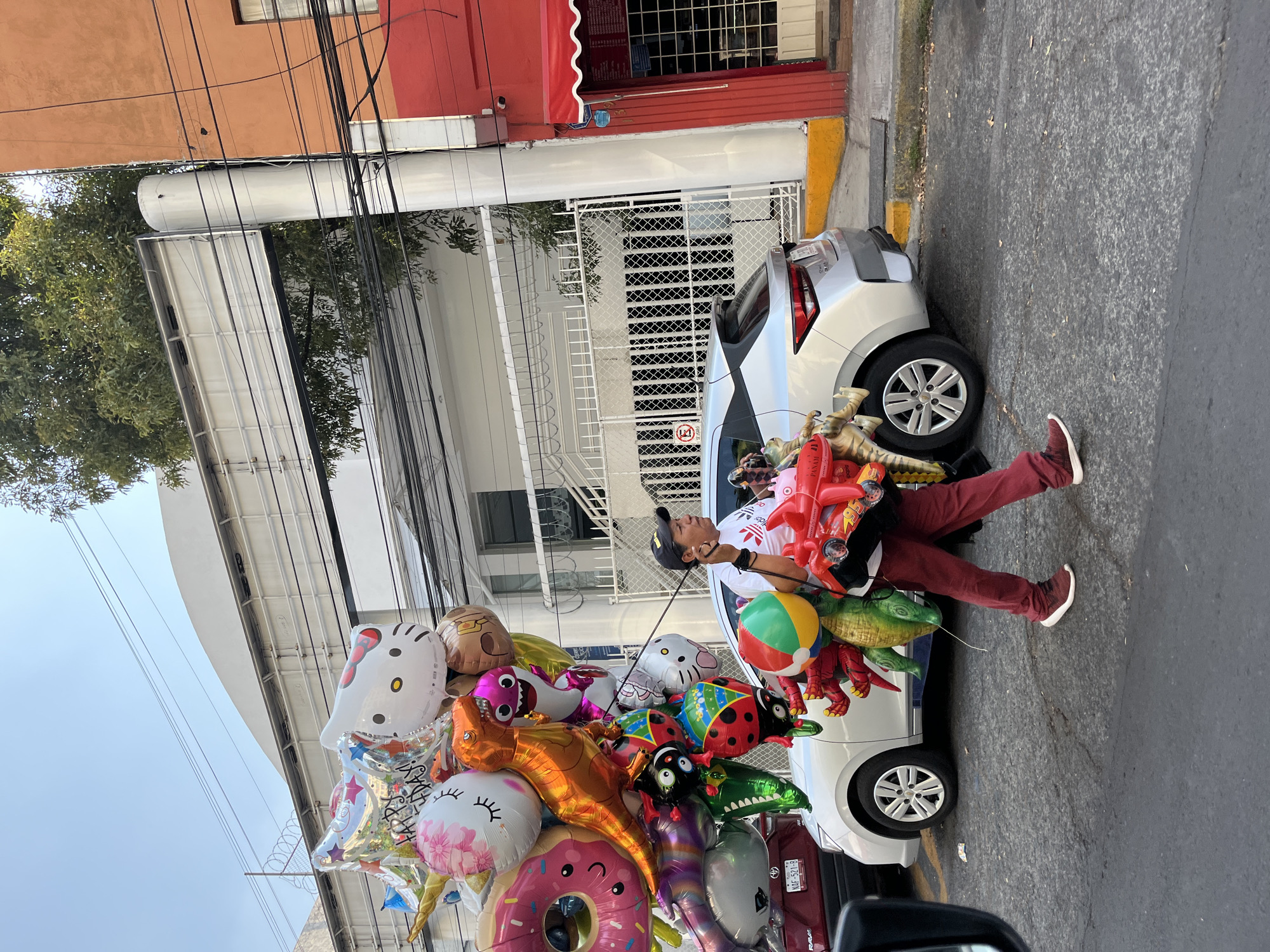 A man stands on the street in Mexico City, Mexico, carrying a large assortment of colorful balloons that are tied to a white sedan parked on the curb. He wears a white t-shirt, maroon pants, maroon shoes, and a black baseball cap. The balloons are of various shapes, including animals, a donut, and a Hello Kitty character. Behind the car is a white building with a wrought iron fence. The facade of the building is painted in a gradient of orange, white, and red, with a red awning above a barred window. Electrical wires are visible above the building. The road appears to be paved and the curb is painted yellow. The scene seems to take place during daylight. Part of another vehicle is visible in the bottom right corner of the image.