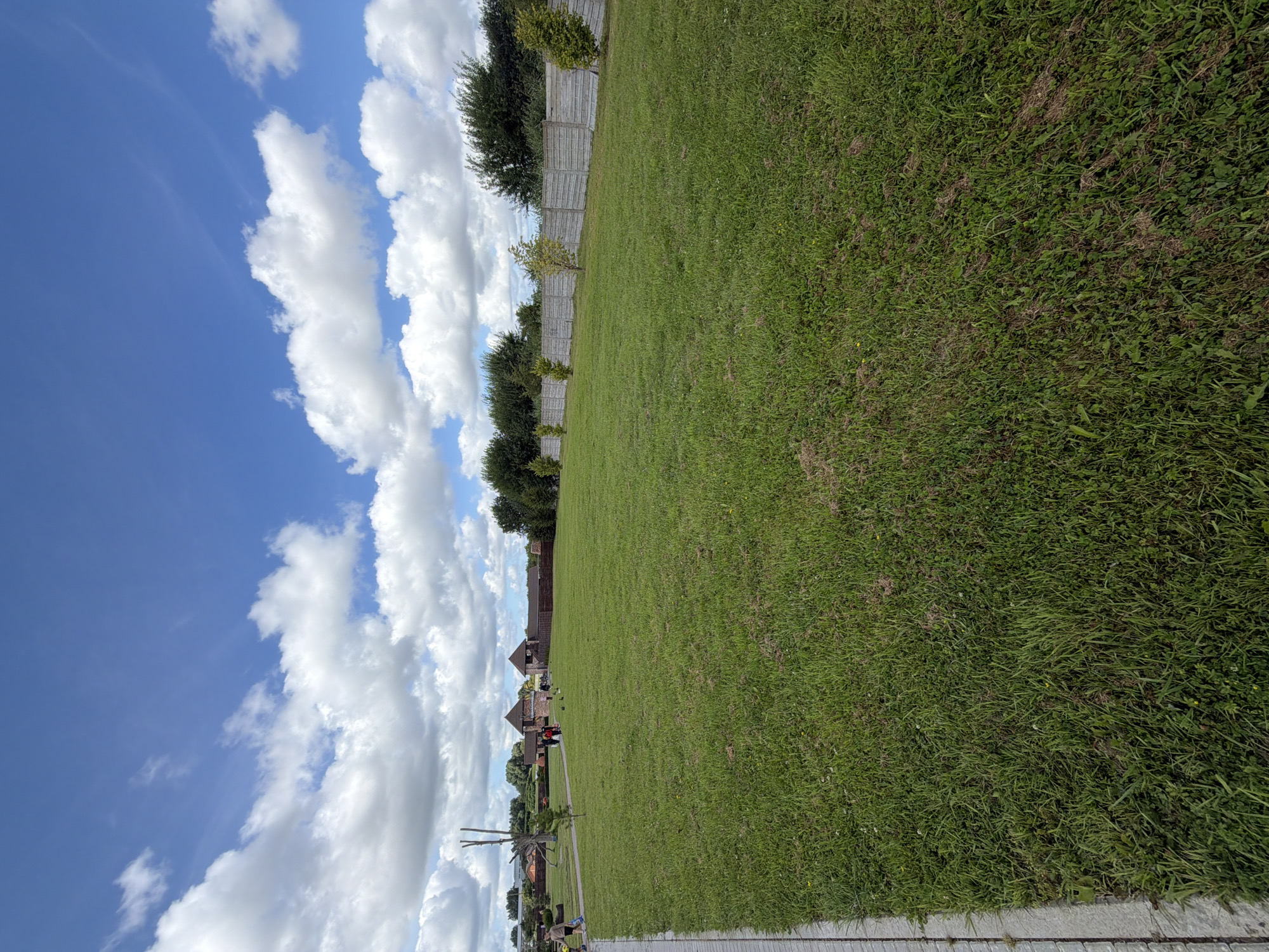 The image depicts a wide, grassy field under a partly cloudy blue sky. A paved walkway runs along the left edge of the frame. In the background, there are trees, a concrete fence, and structures that resemble a medieval-style fortress with towers and wooden walls. Several people are visible walking on the grass and near the structures. The environment appears to be an outdoor recreational area or a park. The location is identified as Konstantinovka, Russia.